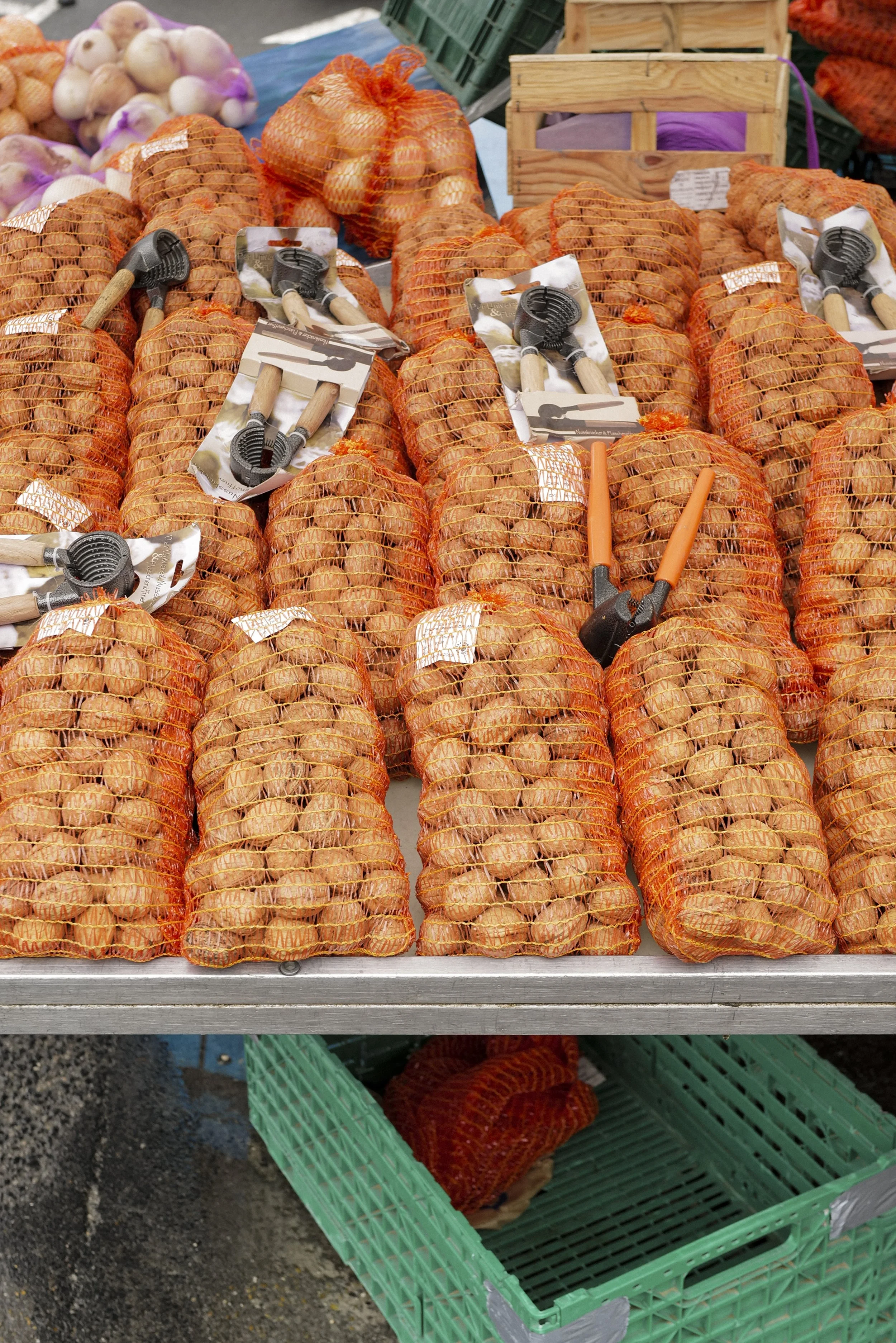 Market stall displaying mesh bags of onions and garlic, some with shovels for sale.