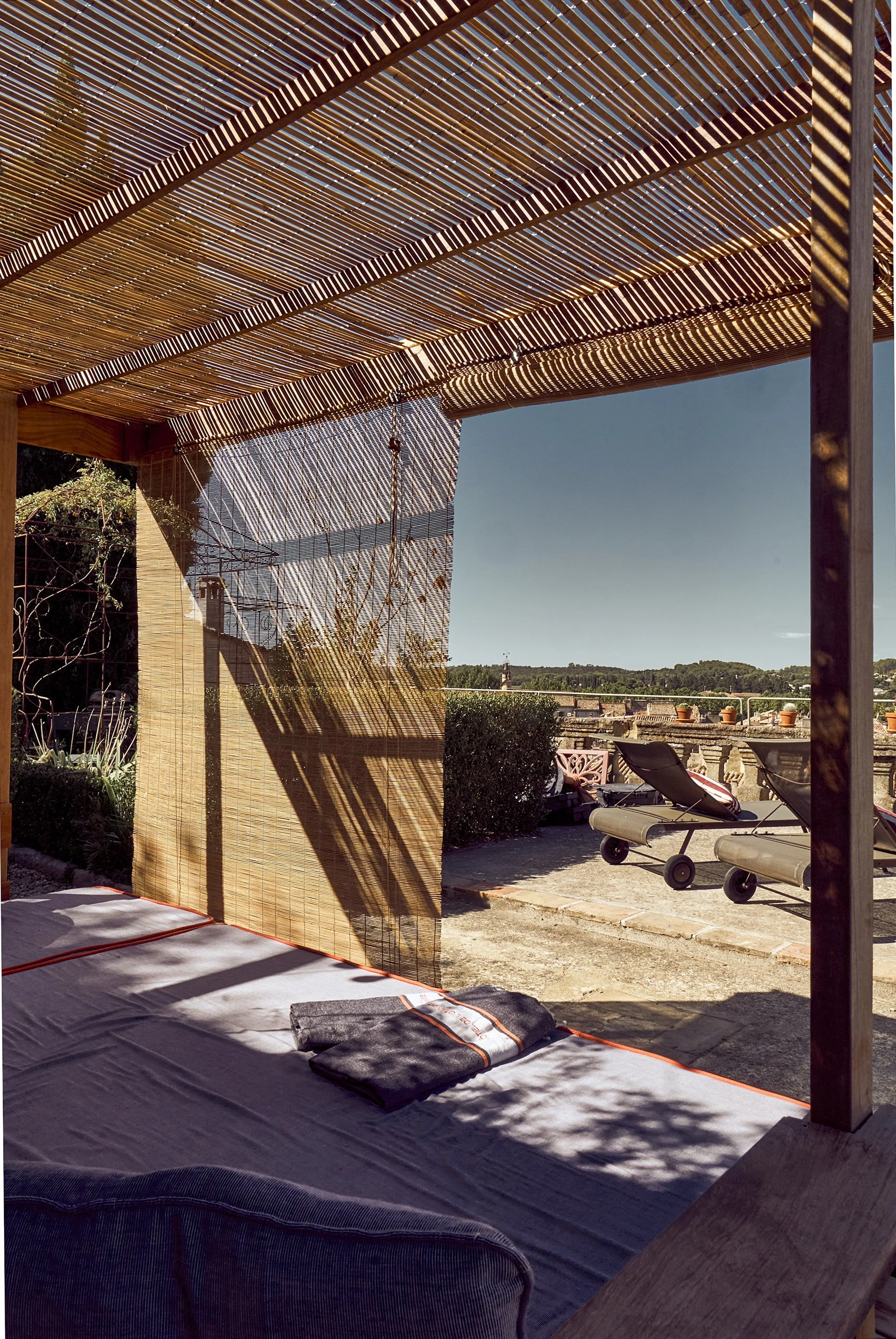 Outdoor patio area with lounge chairs, rolled towels, and a bamboo privacy screen. Sunlight casts shadows through a bamboo roof over this relaxing space, with potted plants and a clear sky in the background.