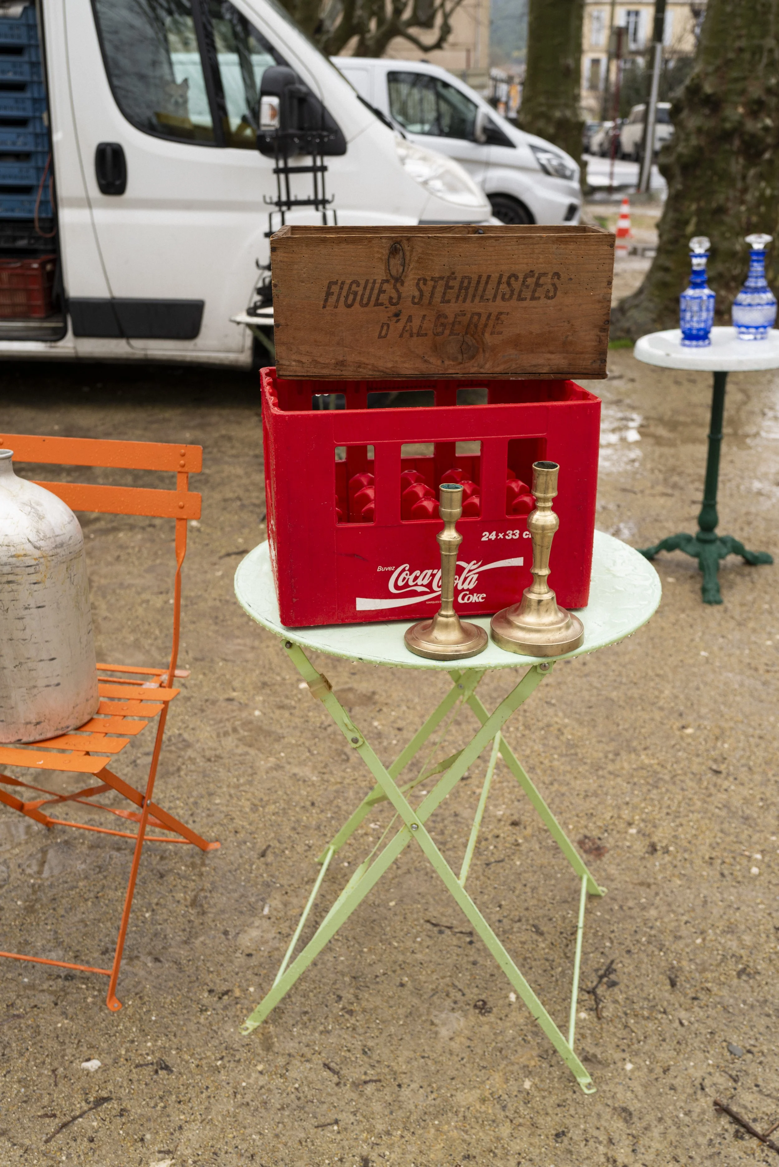 Table with a green folding stand, a Coca-Cola crate holding two brass candle holders, a wooden box with French writing, and two blue glass bottles in the background, outdoors with parked vans and trees.