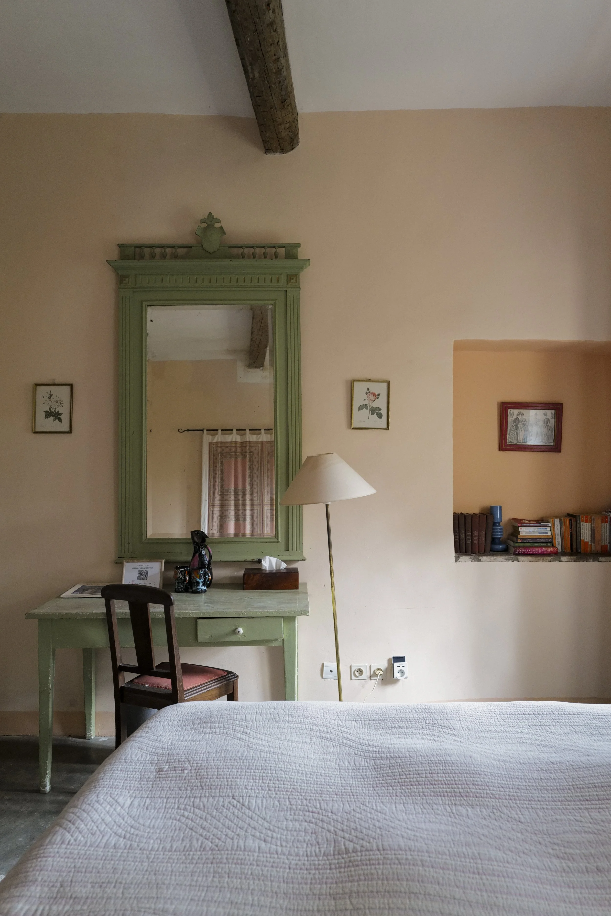A bedroom corner with a light green desk, a dark wooden chair, a mirror, and a white bedside lamp. The wall has framed botanical pictures, and there are electrical outlets and plugs at the bottom. A bed with a white quilt covers part of the scene.