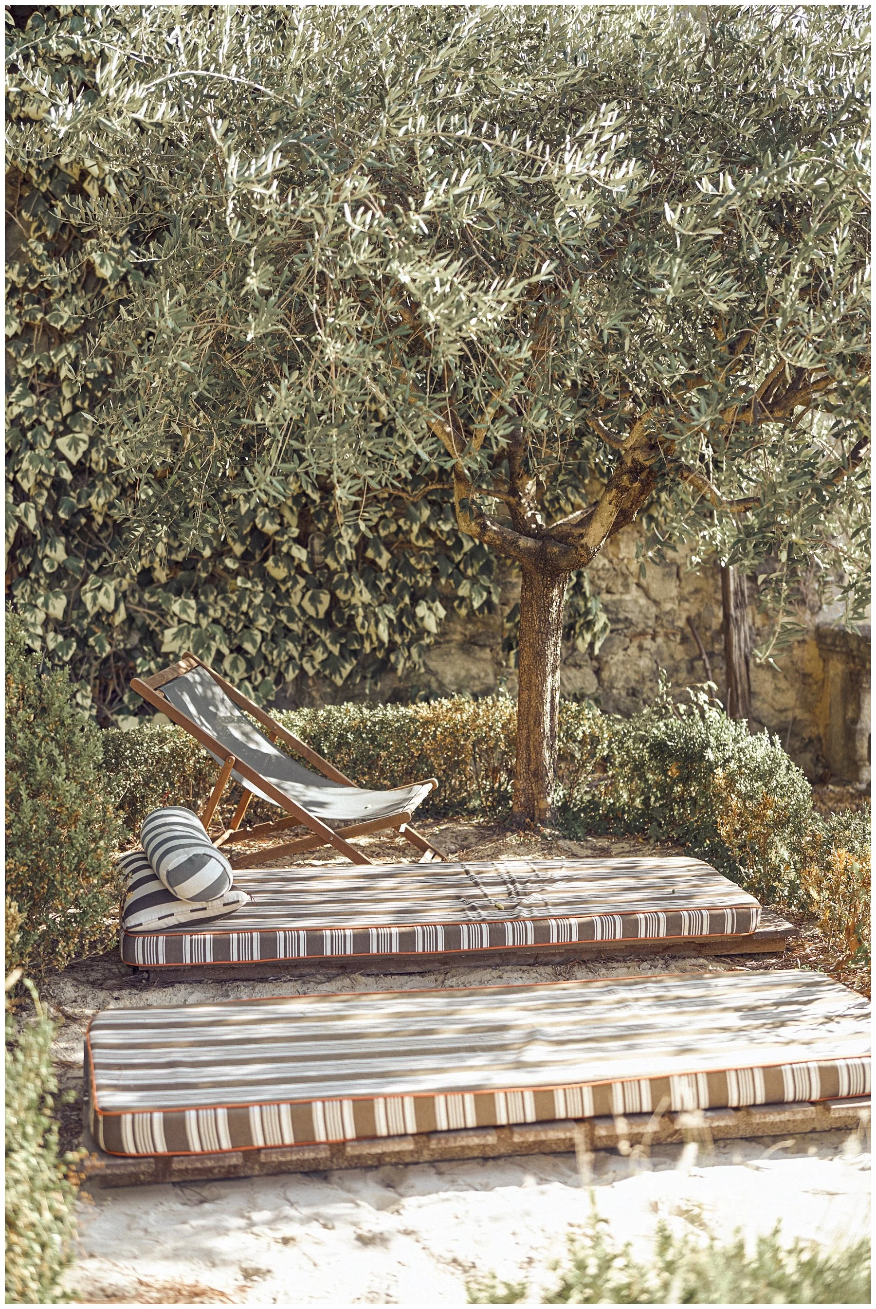 Outdoor garden scene with a large tree, a striped mattress on the ground, two striped pillows, a lounge chair, and lush greenery and bushes.