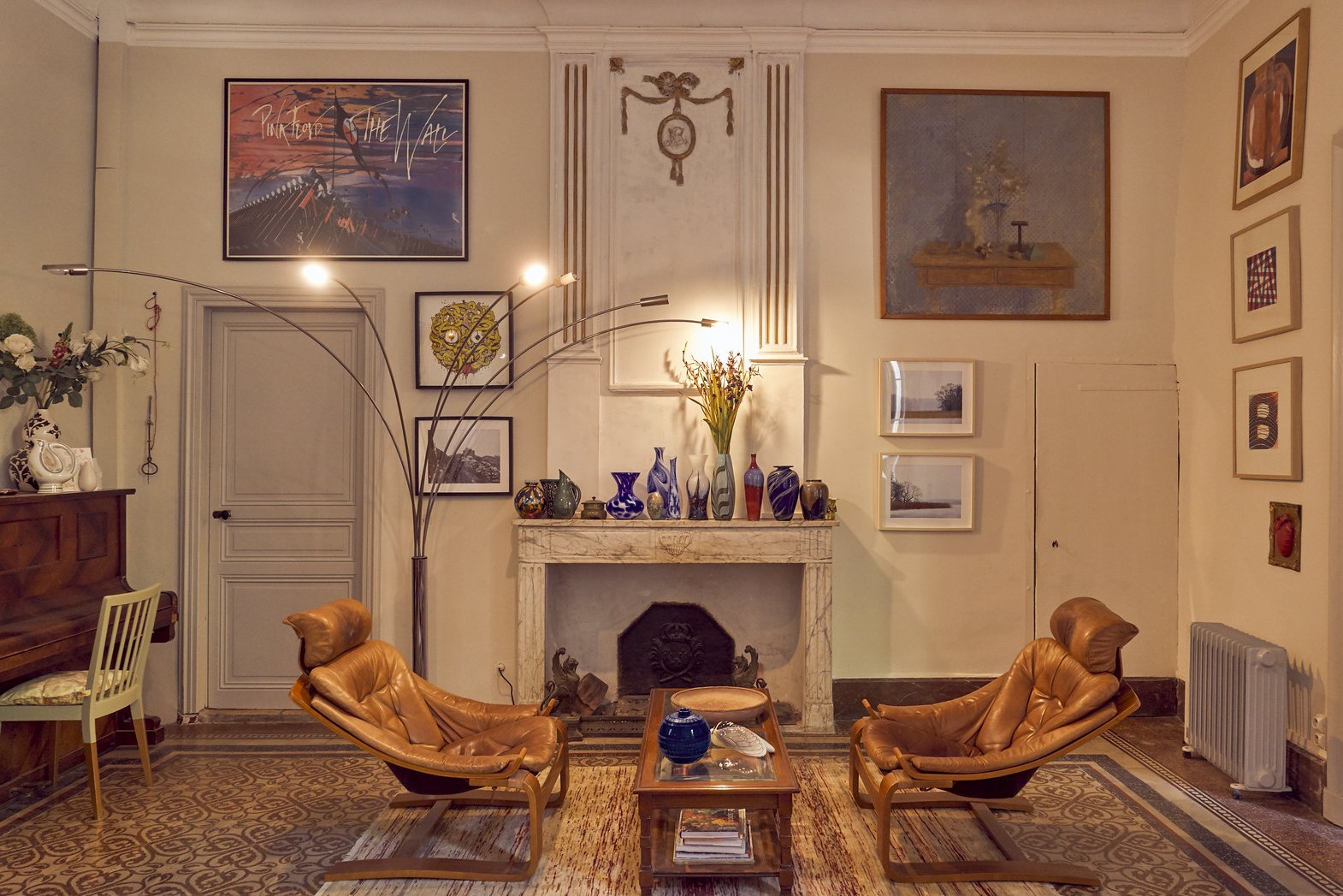 Living room with two tan leather armchairs in front of a fireplace decorated with colorful vases and flowers, artwork on the walls, a tall arched floor lamp, an upright piano with a chair, and a patterned rug.