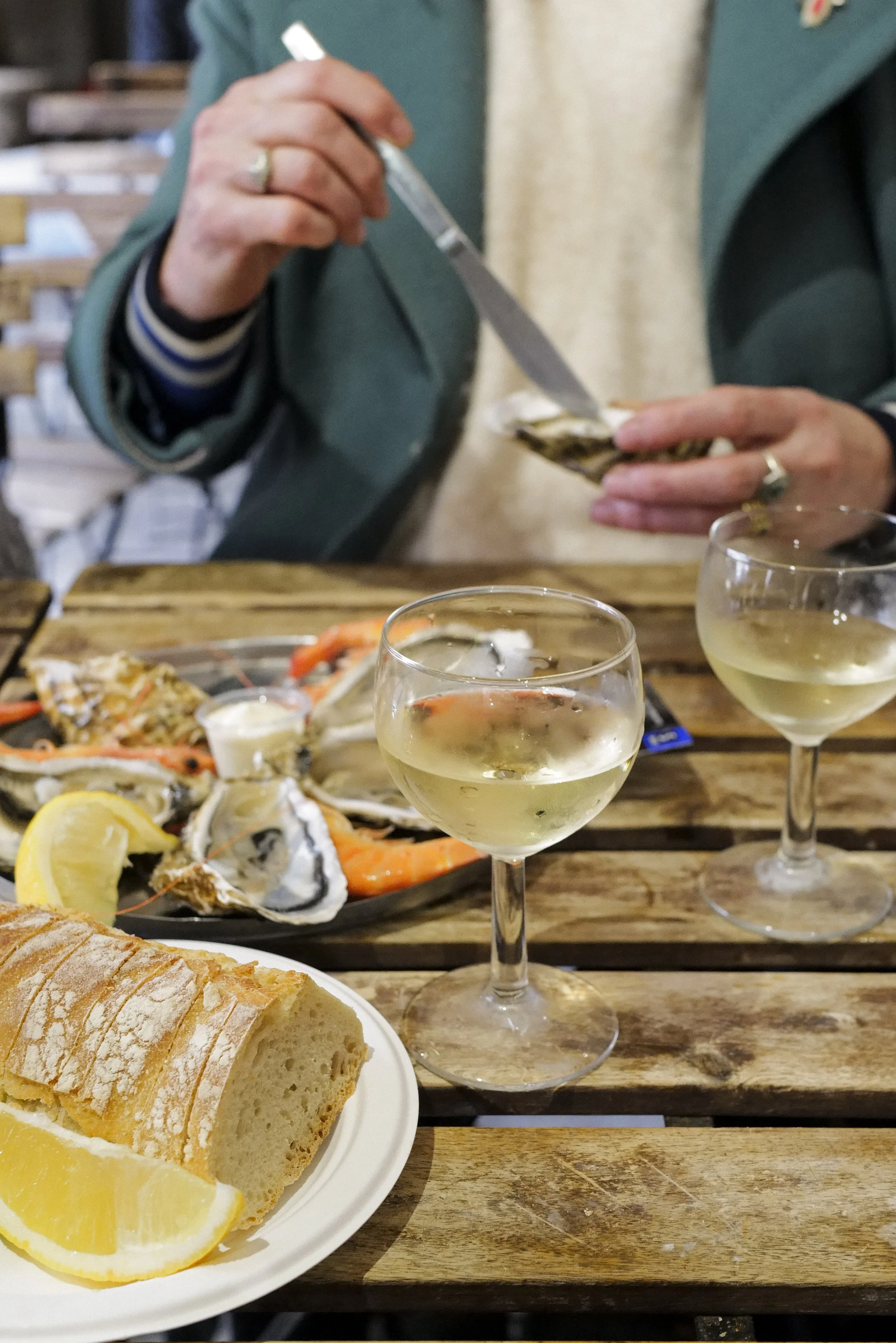 A person spreading butter on a slice of bread with oysters, shrimp, lemon, and cheese spread on a wooden table, along with two glasses of white wine.