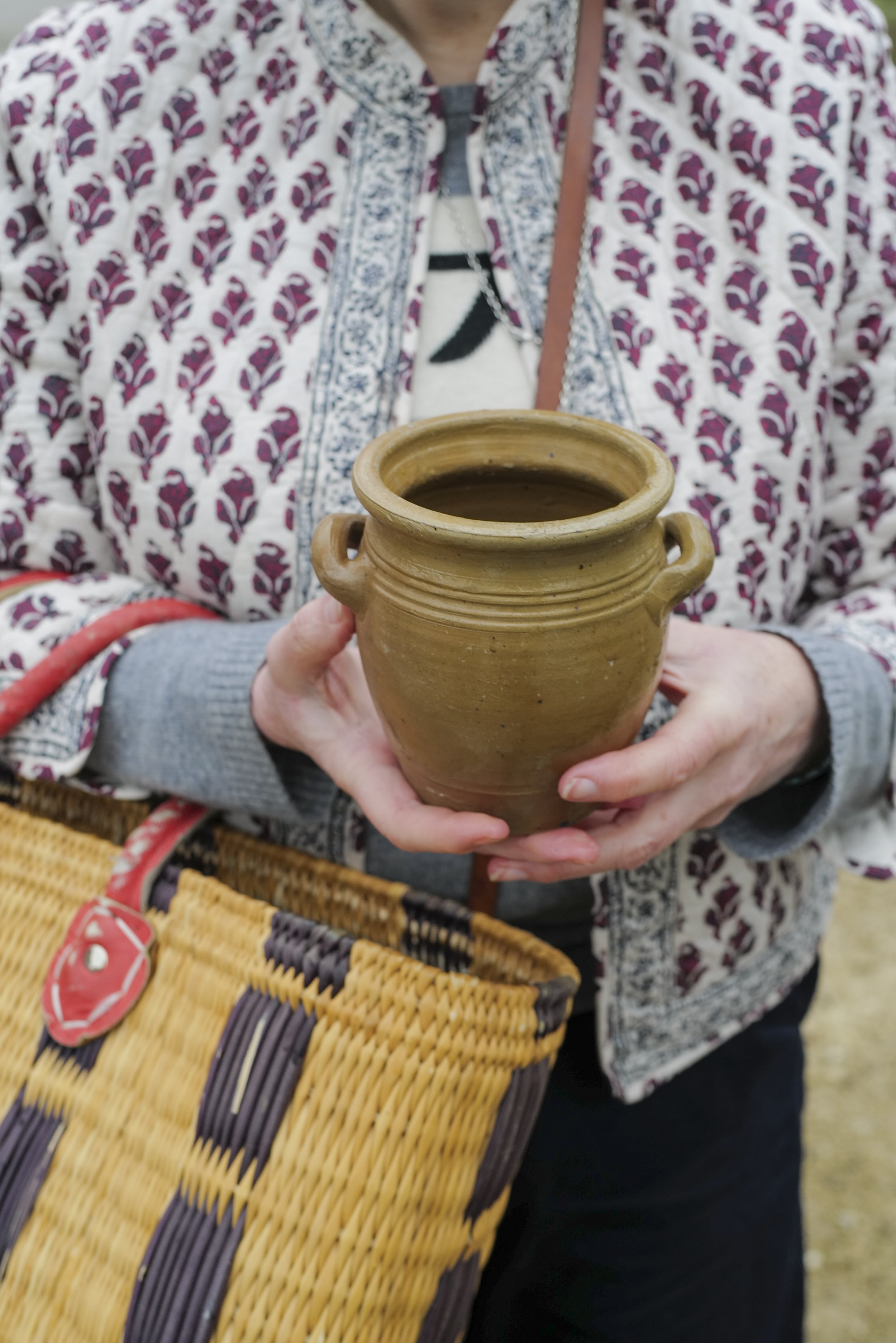 Person holding a brown ceramic mug filled with a dark beverage, wearing a patterned fabric jacket and gray sweater, with a woven basket nearby.