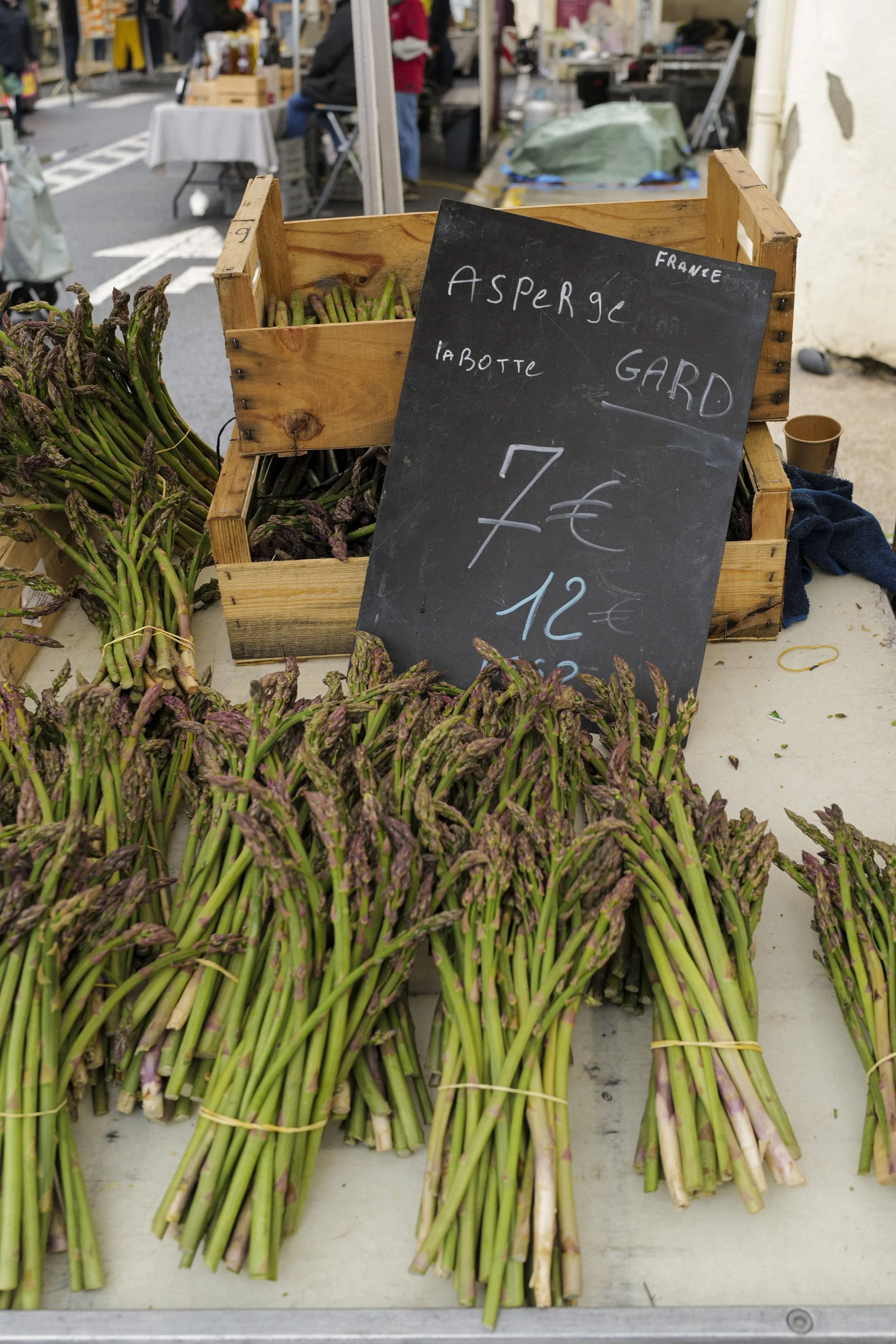 Fresh bunches of asparagus on a market table with a black chalkboard sign listing the price in euros, located at an outdoor market with other stalls and shoppers in the background.