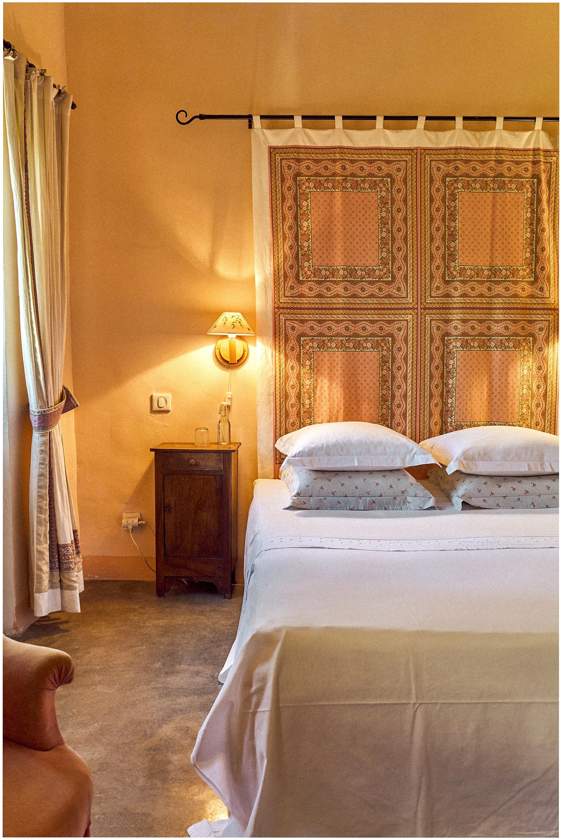 A cozy bedroom with a large bed covered in white linens and decorative pillows. Behind the bed is a decorative fabric headboard with intricate patterns. To the left, a small wooden nightstand with a glass bottle, a glass, and a wall-mounted lamp prov