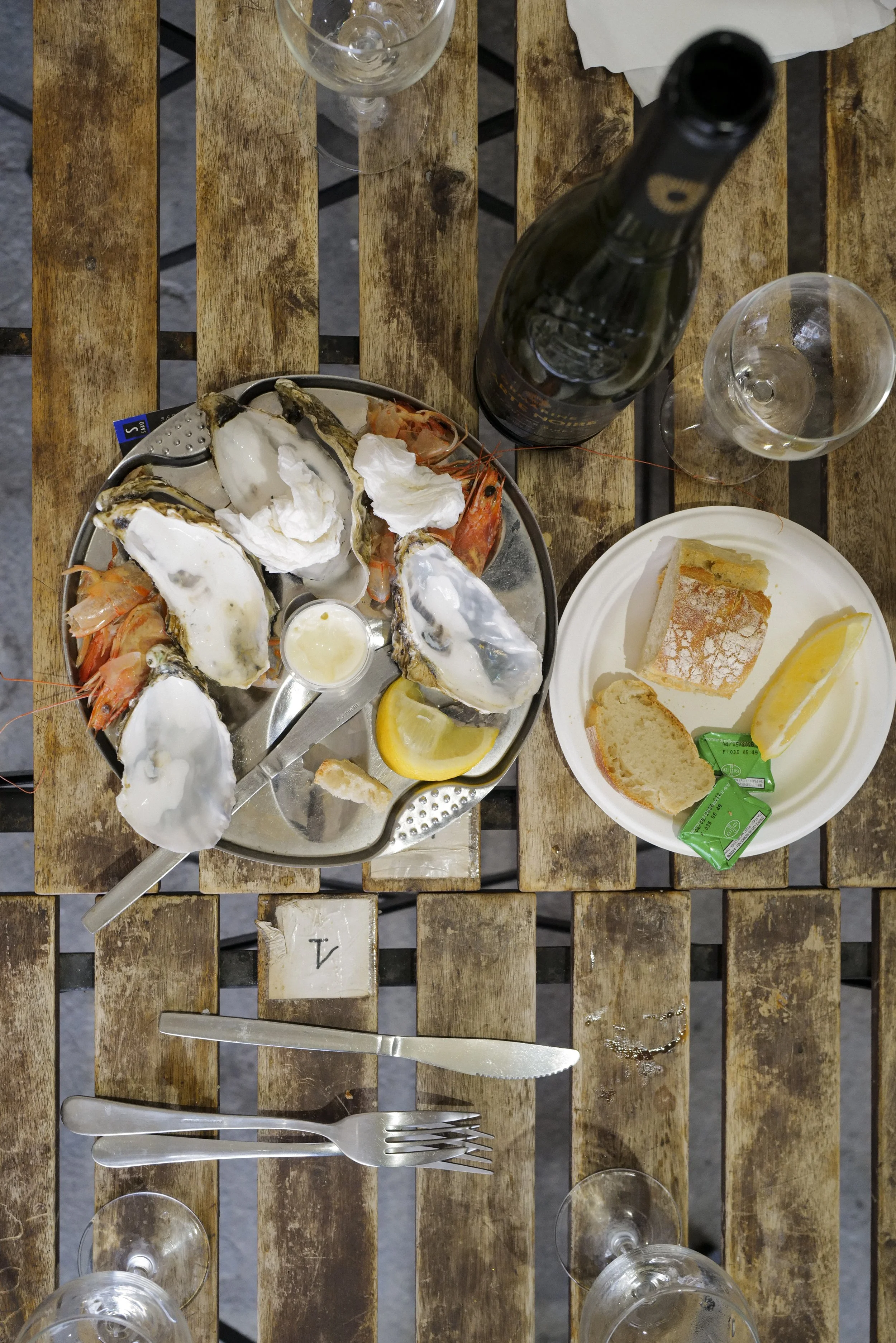 A table with a plate of oysters topped with white sauce, shrimp, lemon wedges, a bottle of wine, empty wine glasses, and utensils on a wooden outdoor table.