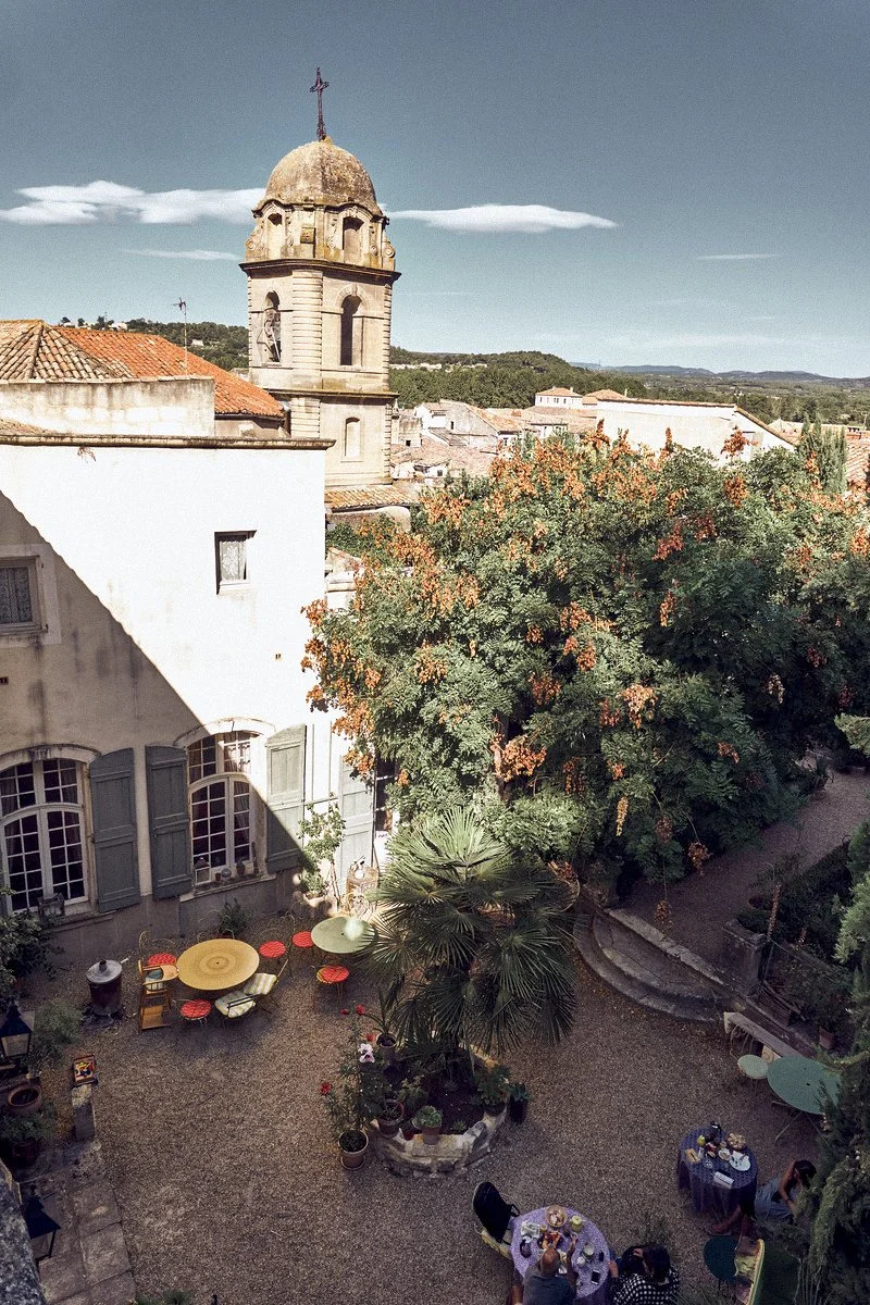 A cozy courtyard with outdoor seating, large trees with orange flowers, and historic buildings with a church tower in the background under a clear blue sky.