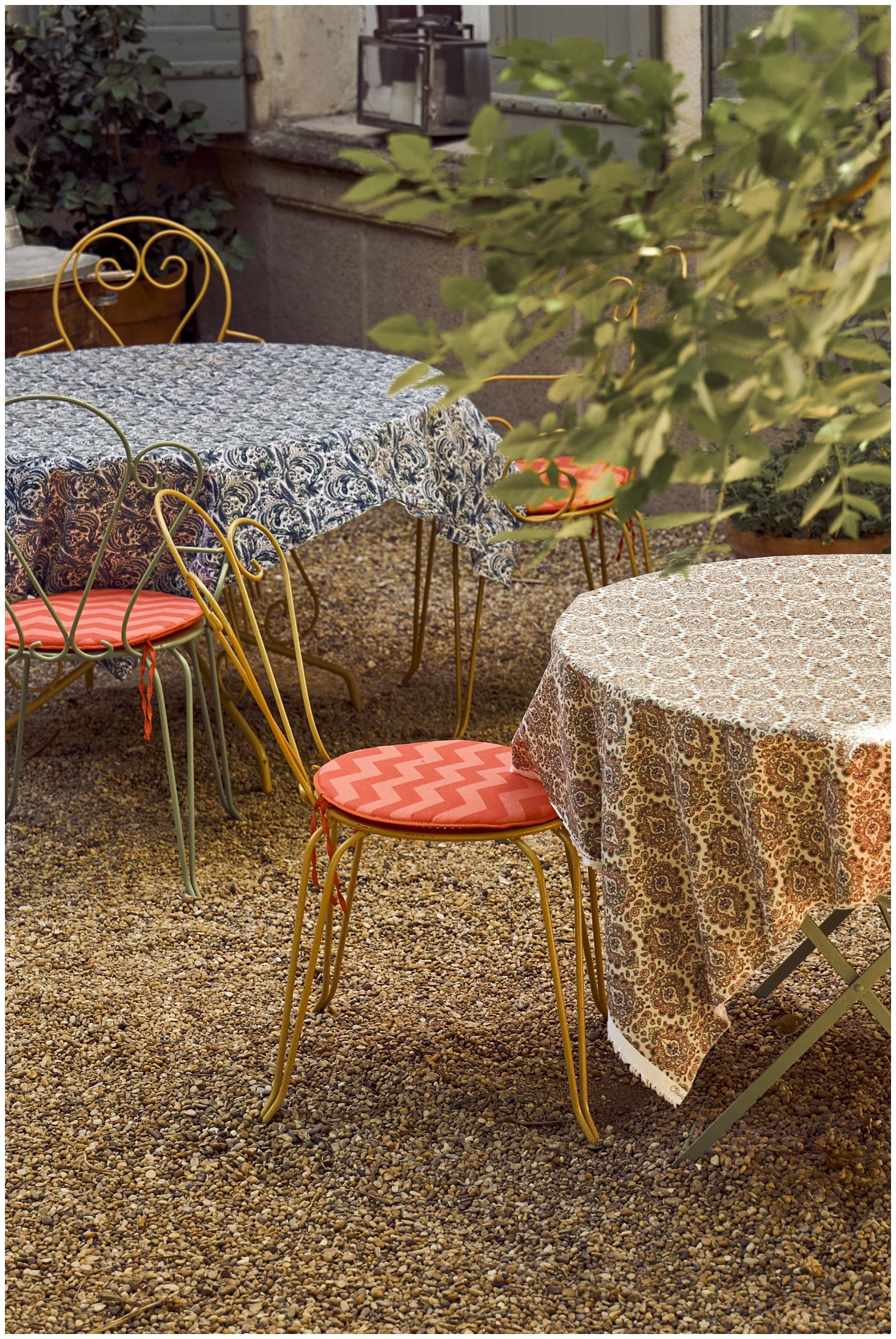 Outdoor patio with two round tables covered with patterned tablecloths, and yellow metal chairs with pink cushions. Pebble ground, greenery, and a stone wall with a window and a small metal lantern in the background.