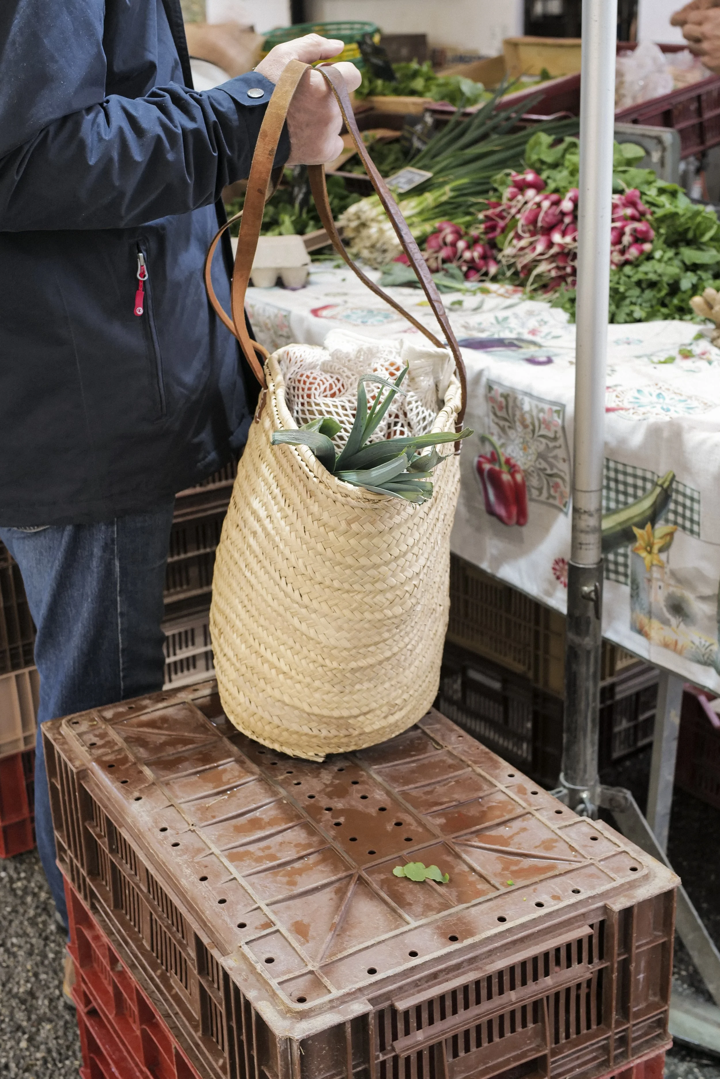 A person shopping at an outdoor market, holding a woven basket with green onions inside. The background has tables with various fresh vegetables, including radishes and leafy greens, and crates for displaying produce.