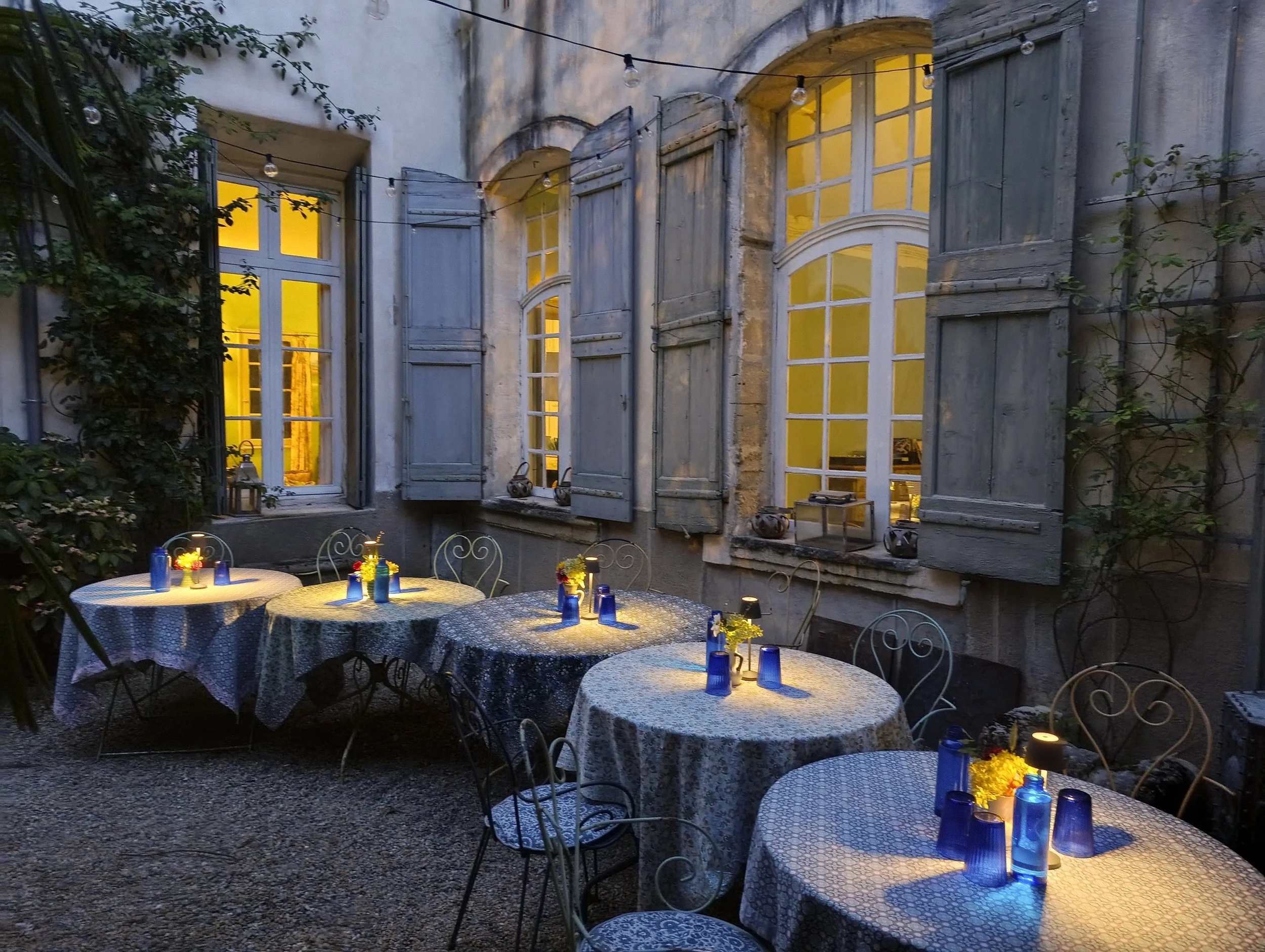 Outdoor patio with tables covered in patterned tablecloths, lit by candles and blue vases, against a weathered building with large yellow-lit windows and open wooden shutters, decorated with string lights and greenery.