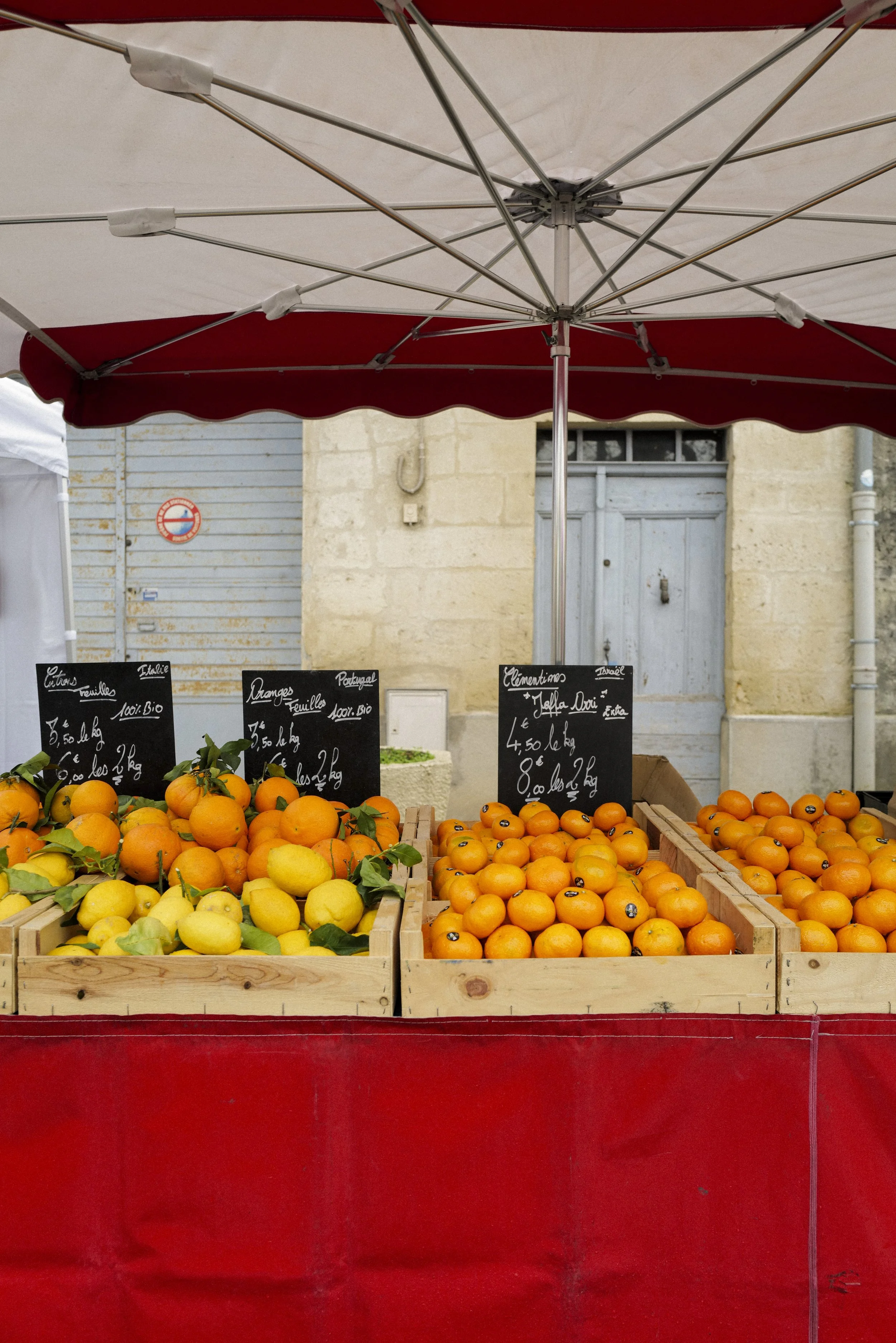 A street market stand with crates of yellow and orange citrus fruits, under a red and white striped umbrella, in front of an old building with a closed blue door and shuttered windows.