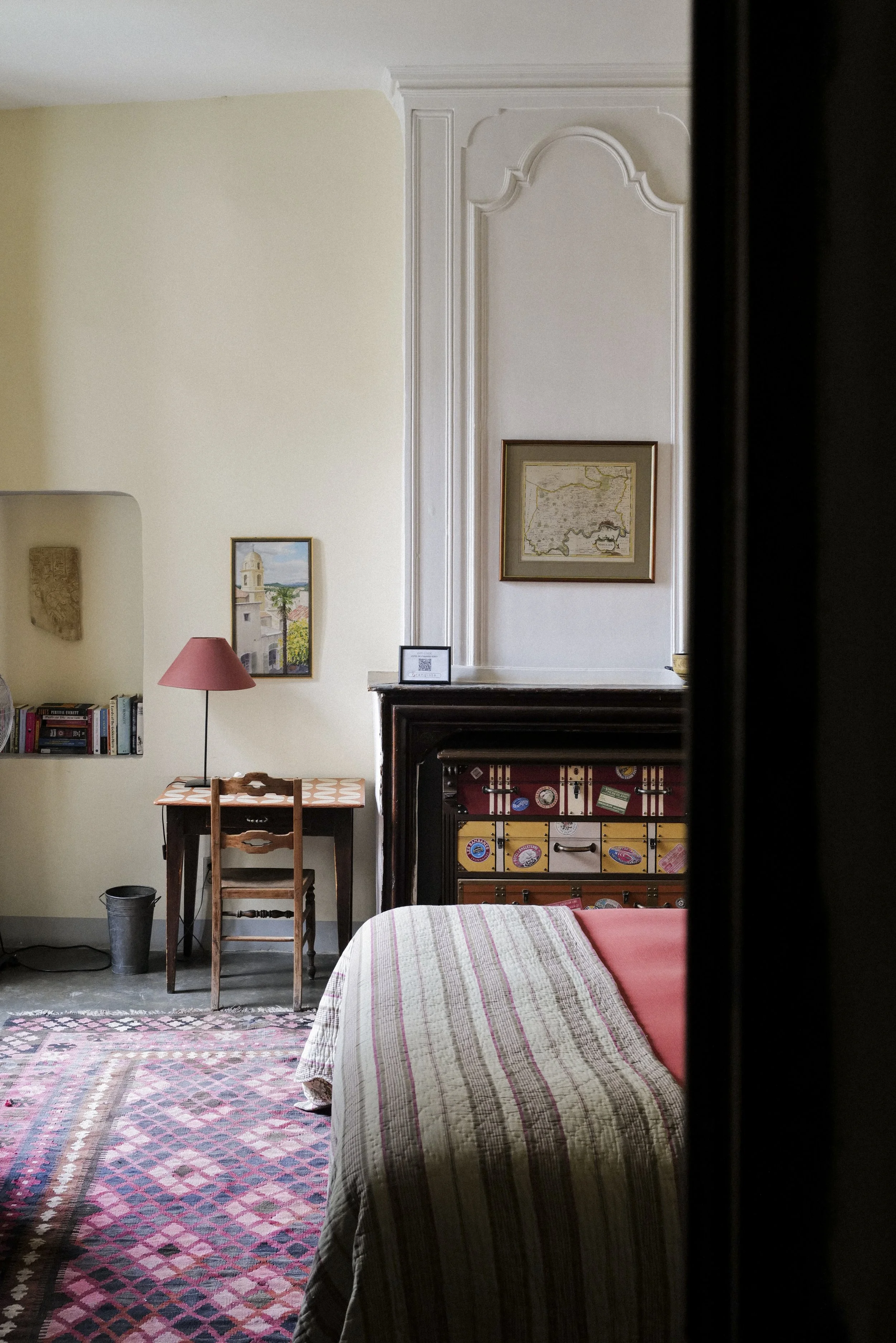 A corner of a room with a beige wall, a framed painting of a European cityscape, a small desk with a wooden chair, a pink lampshade, a shelf with books, a framed photograph, and a fireplace with a framed map above it. Part of a bed with a multicolore