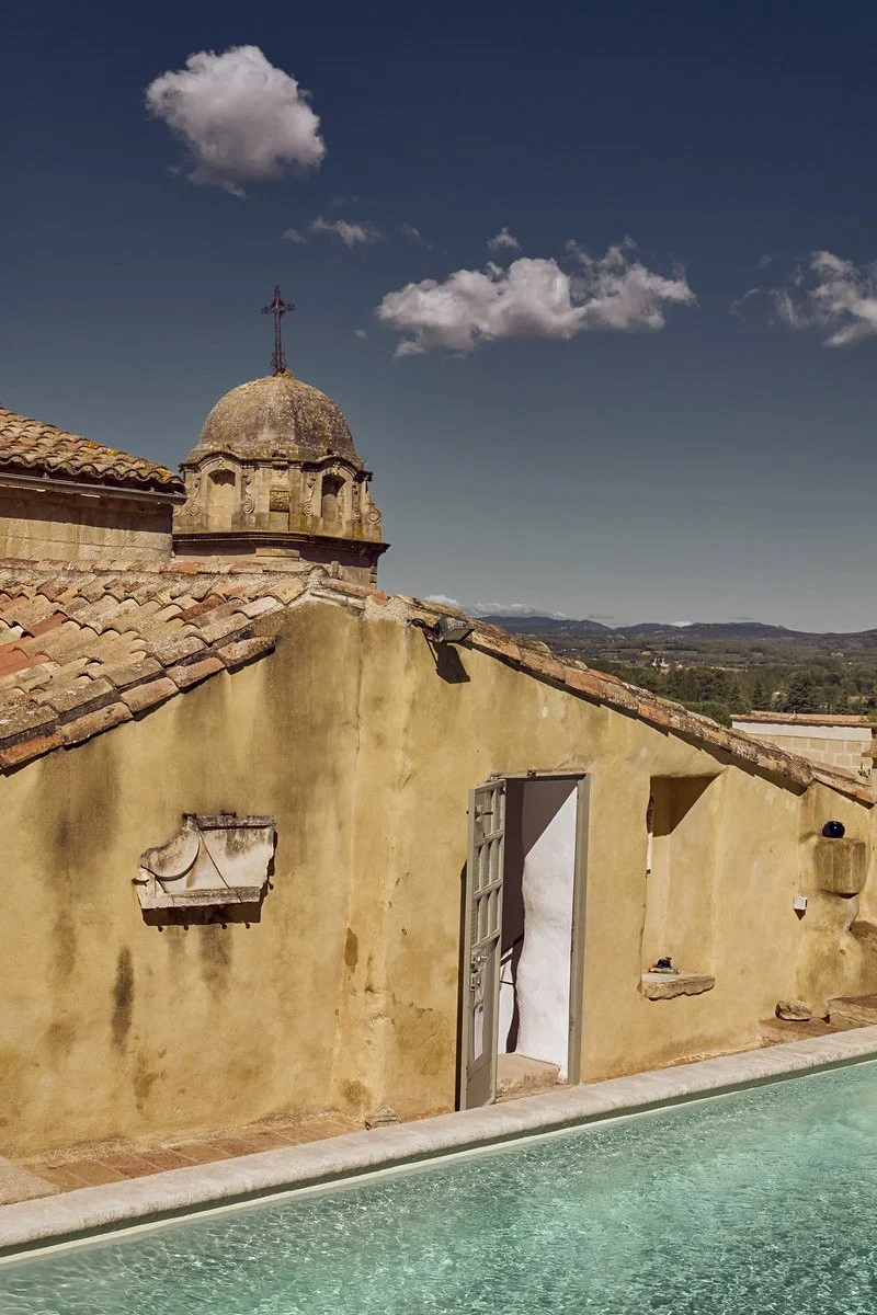 A rustic yellow building with a tiled roof, a small courtyard with a swimming pool, and a church dome with an iron cross on top under a blue sky with a few white clouds.
