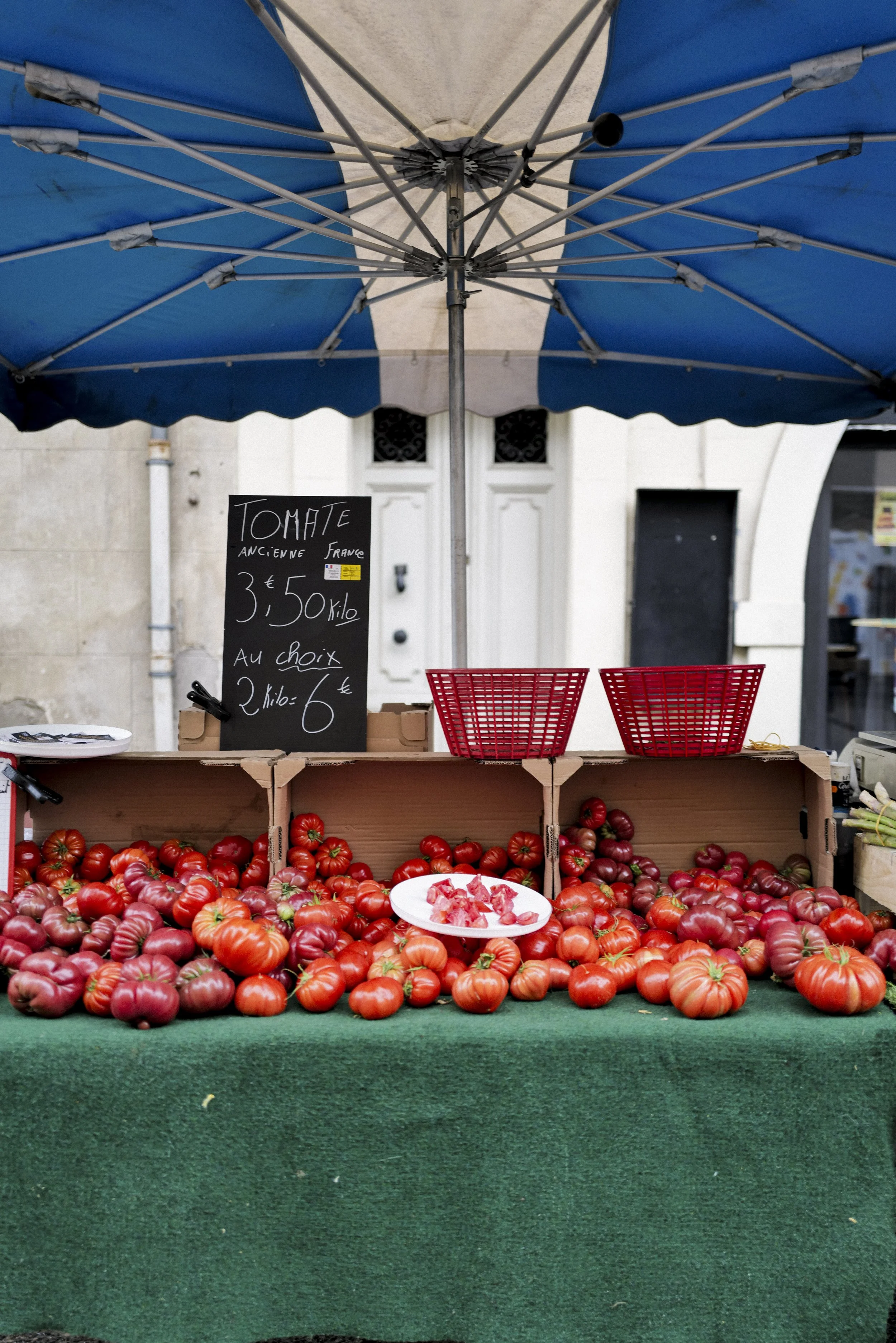 Market stall with fresh tomatoes on display under a large blue and white umbrella, with a chalkboard sign listing prices and options.
