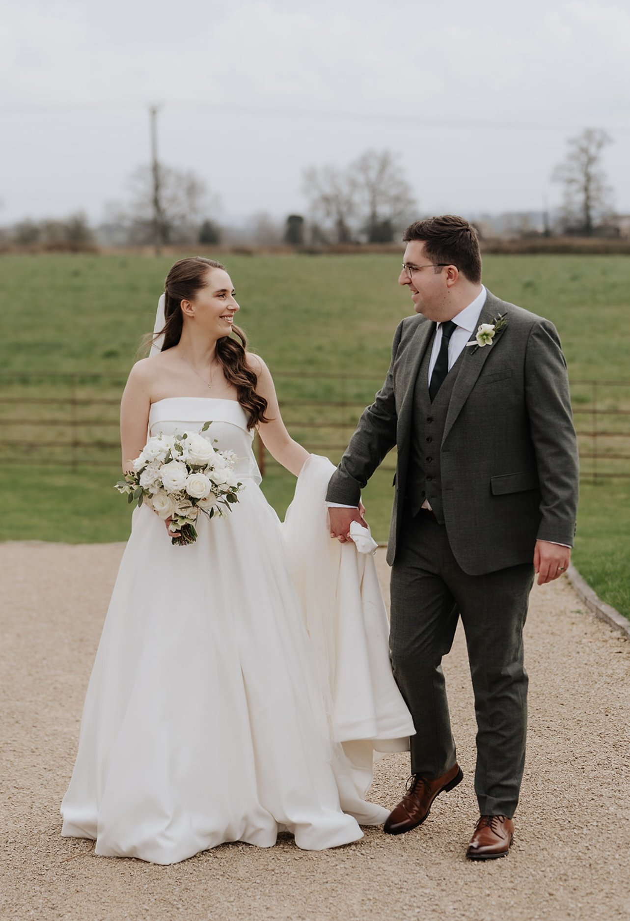 A bride in a white wedding dress holds a bouquet of white flowers while walking with a groom in a gray suit on a gravel path with green fields in the background.