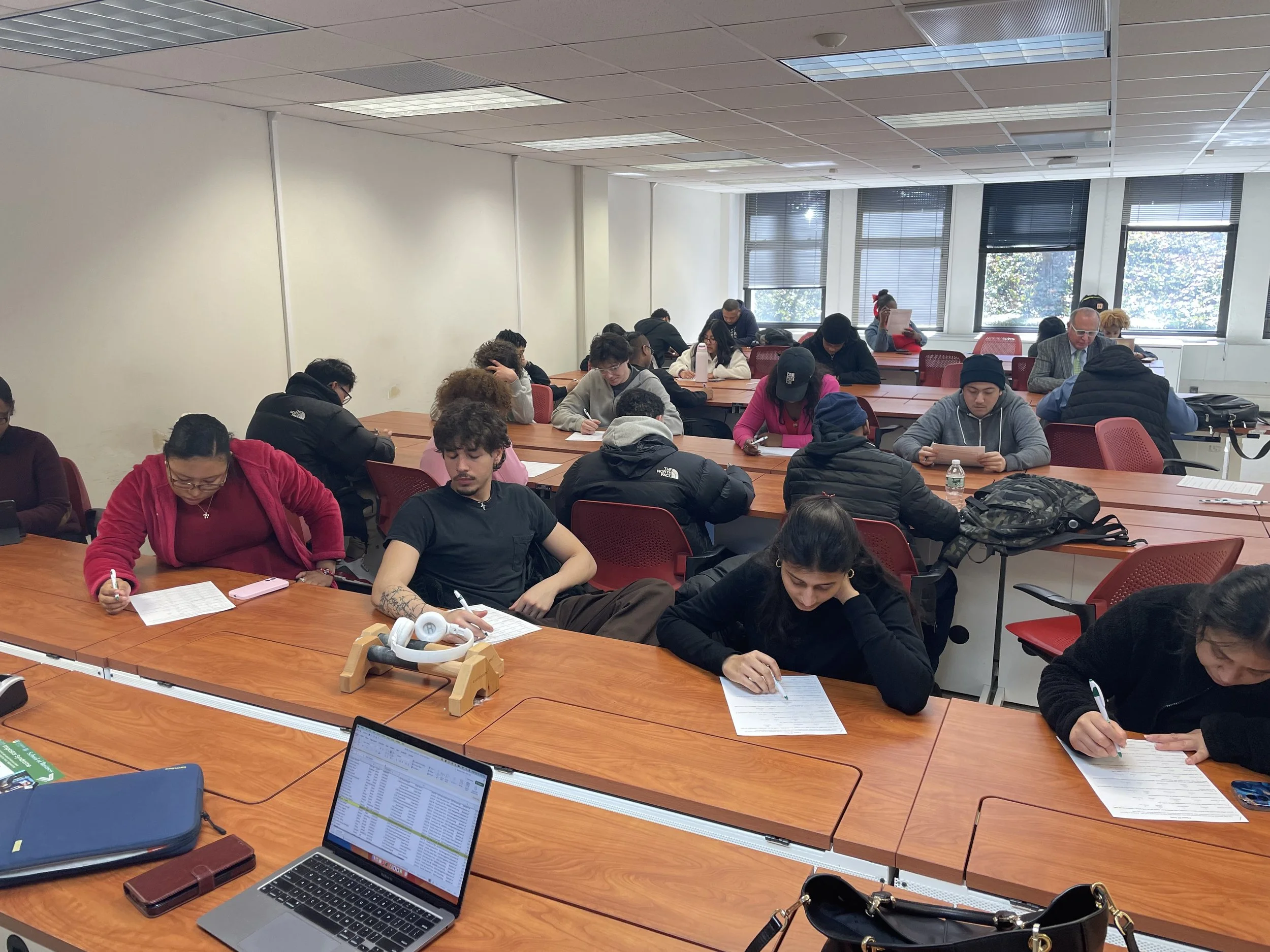 A classroom full of students sitting at desks taking notes, with computer laptops open in front of some students, windows behind them showing trees outside, and a white wall on the side.