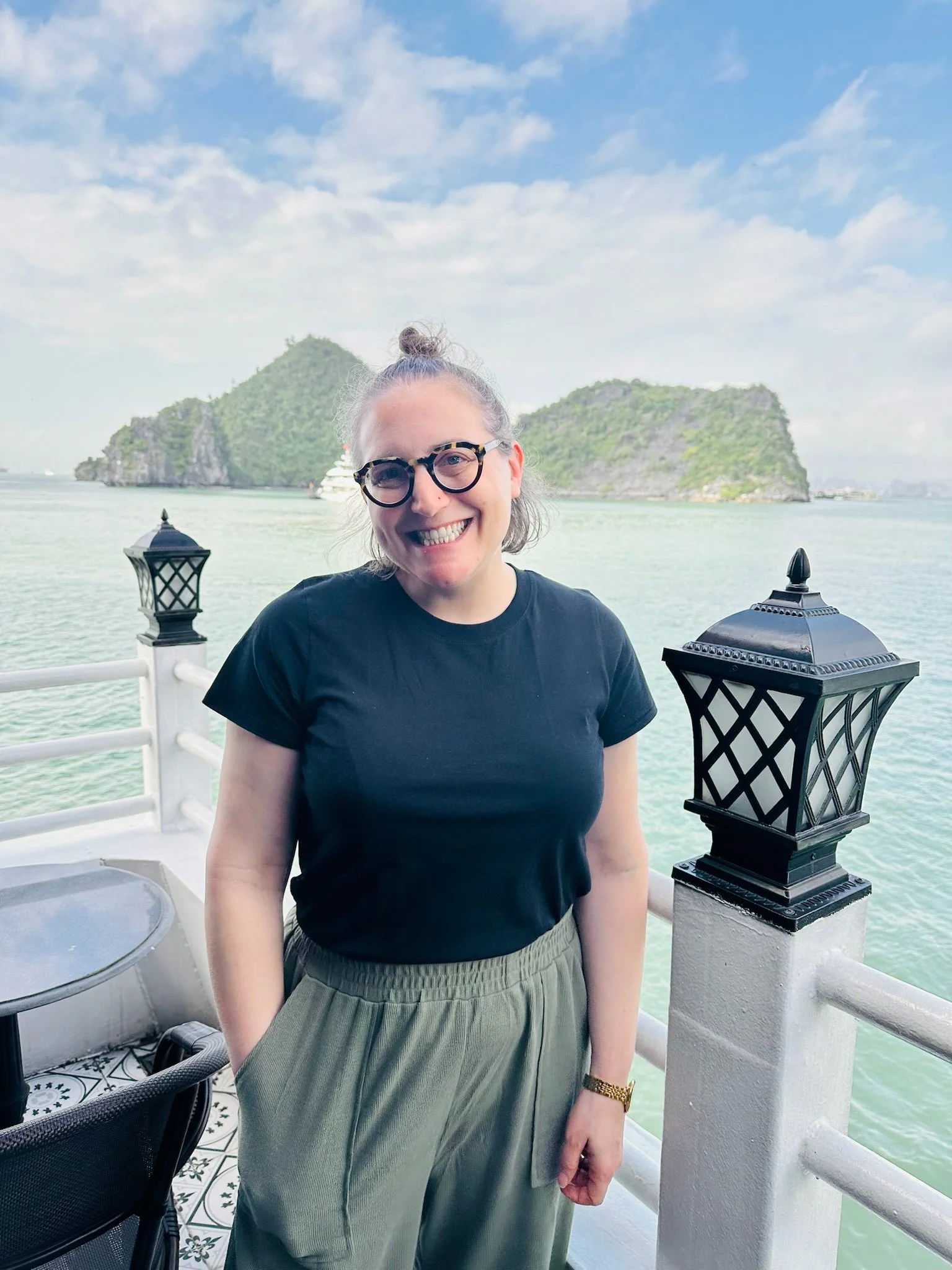 A woman, wearing glasses, a black T-shirt, and green pants, standing on a boat with water, islands, and a yacht in the background under a cloudy sky in Vietnam.