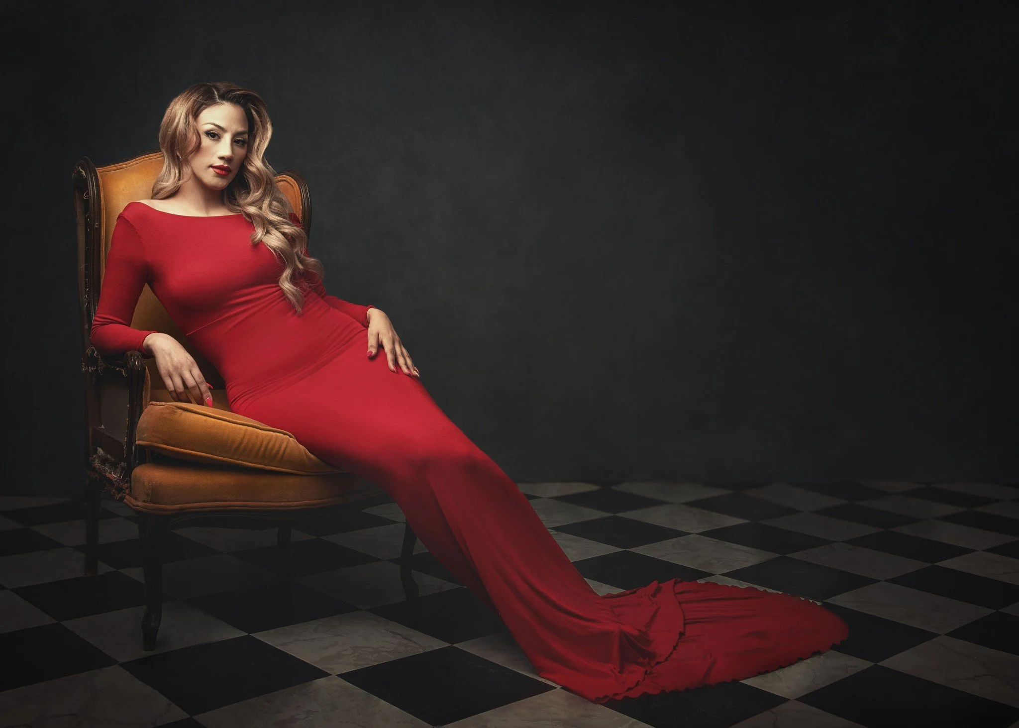 A woman in a long red dress reclining on a vintage-style armchair against a dark background with a black and white checkered floor.