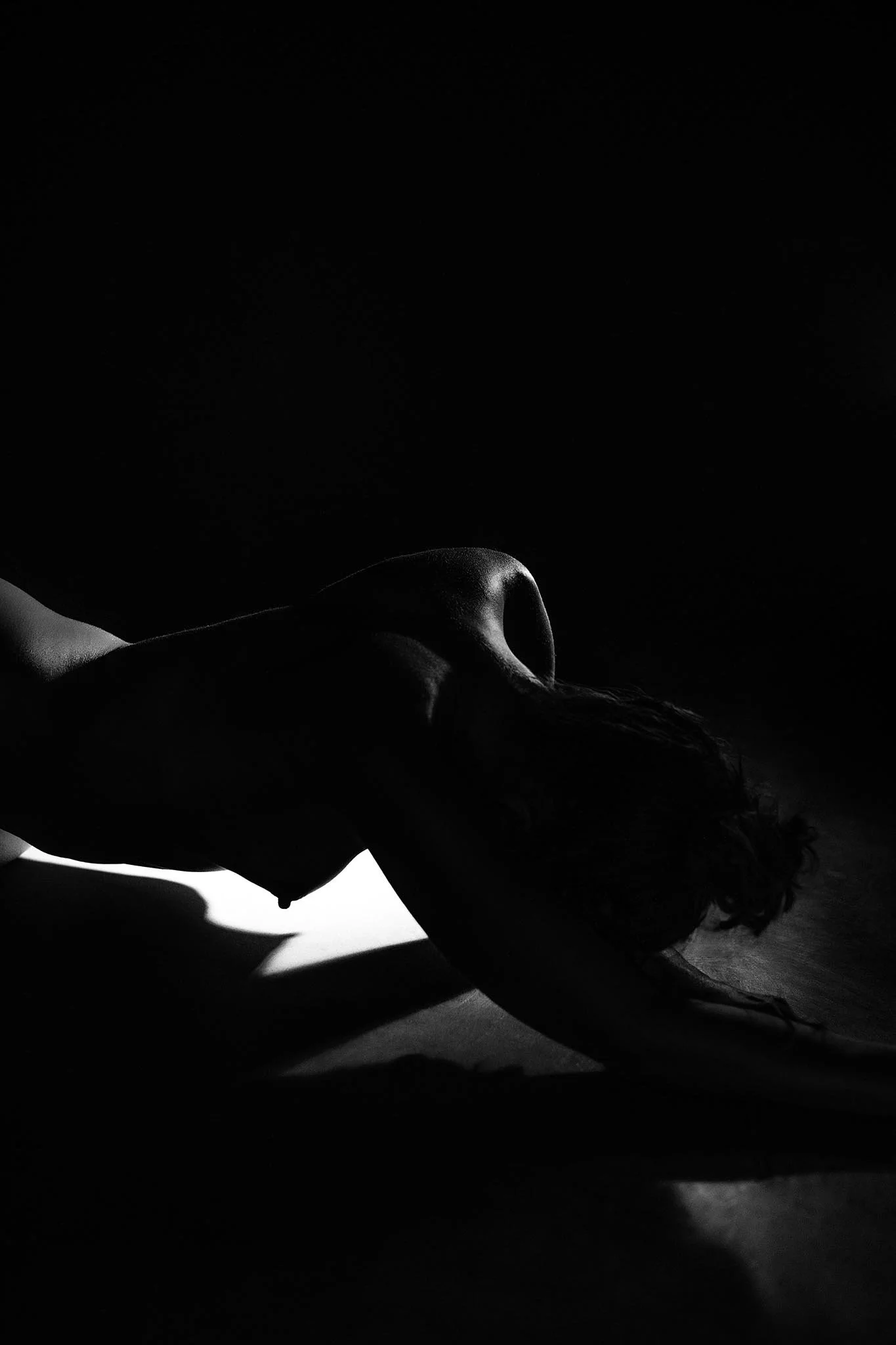 A black-and-white silhouette of a person with long hair, kneeling and leaning forward on a dark surface, illuminated by a single light source.