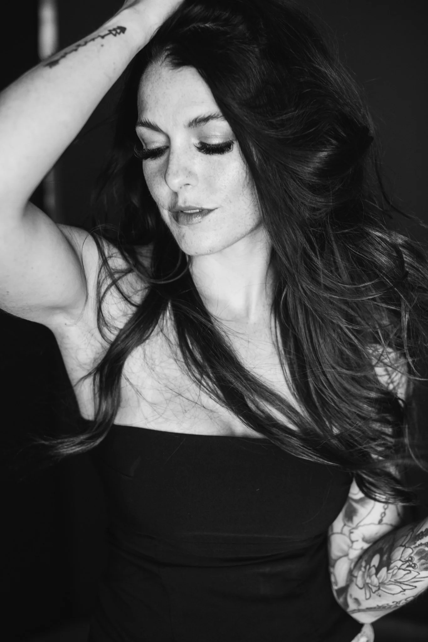 A black-and-white portrait of a young woman with long, dark hair, closed eyes, and makeup, touching her hair with one hand, wearing a strapless top, with a tattoo on her arm.