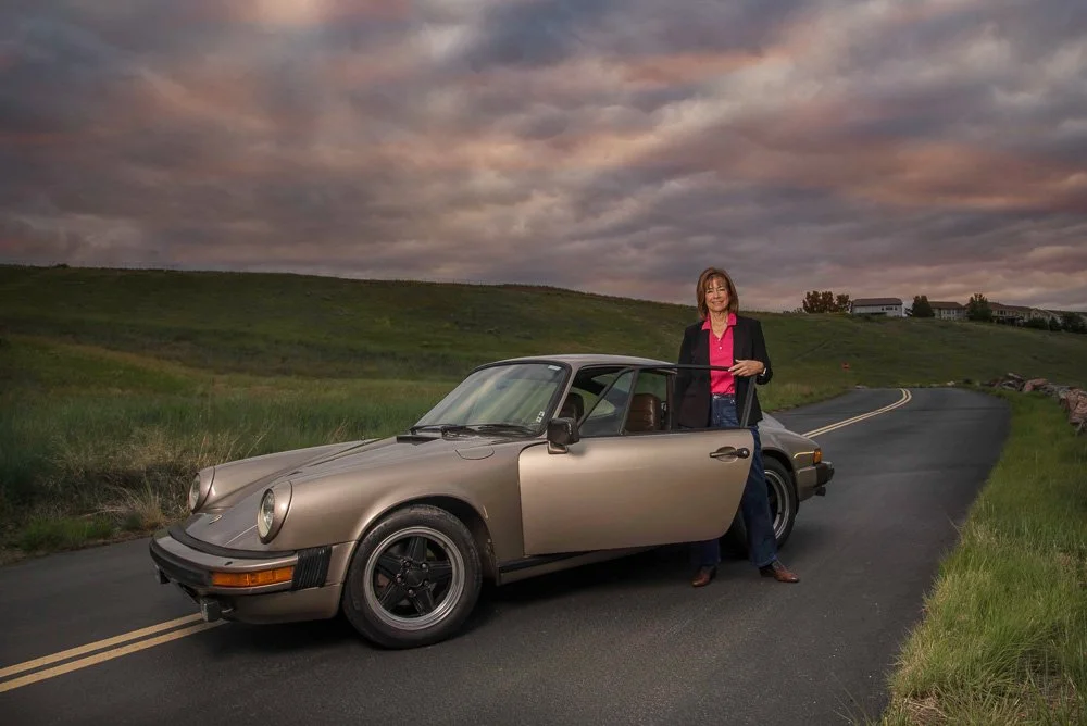 A woman standing next to a vintage beige Porsche 911 on a winding rural road at sunset, with grassy fields and hills in the background.