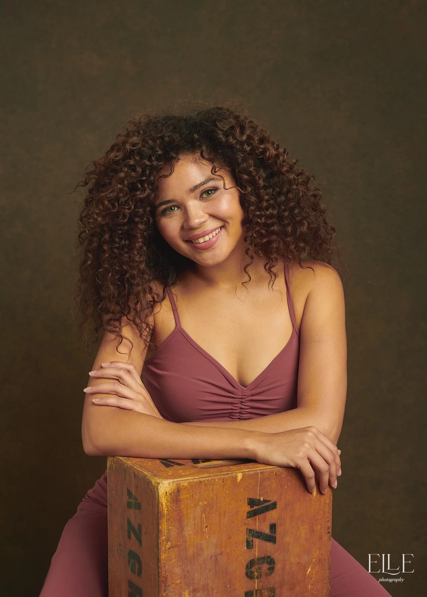 A smiling woman with curly brown hair, in a mauve dress, leaning on her arms on a wooden crate against a dark studio background.