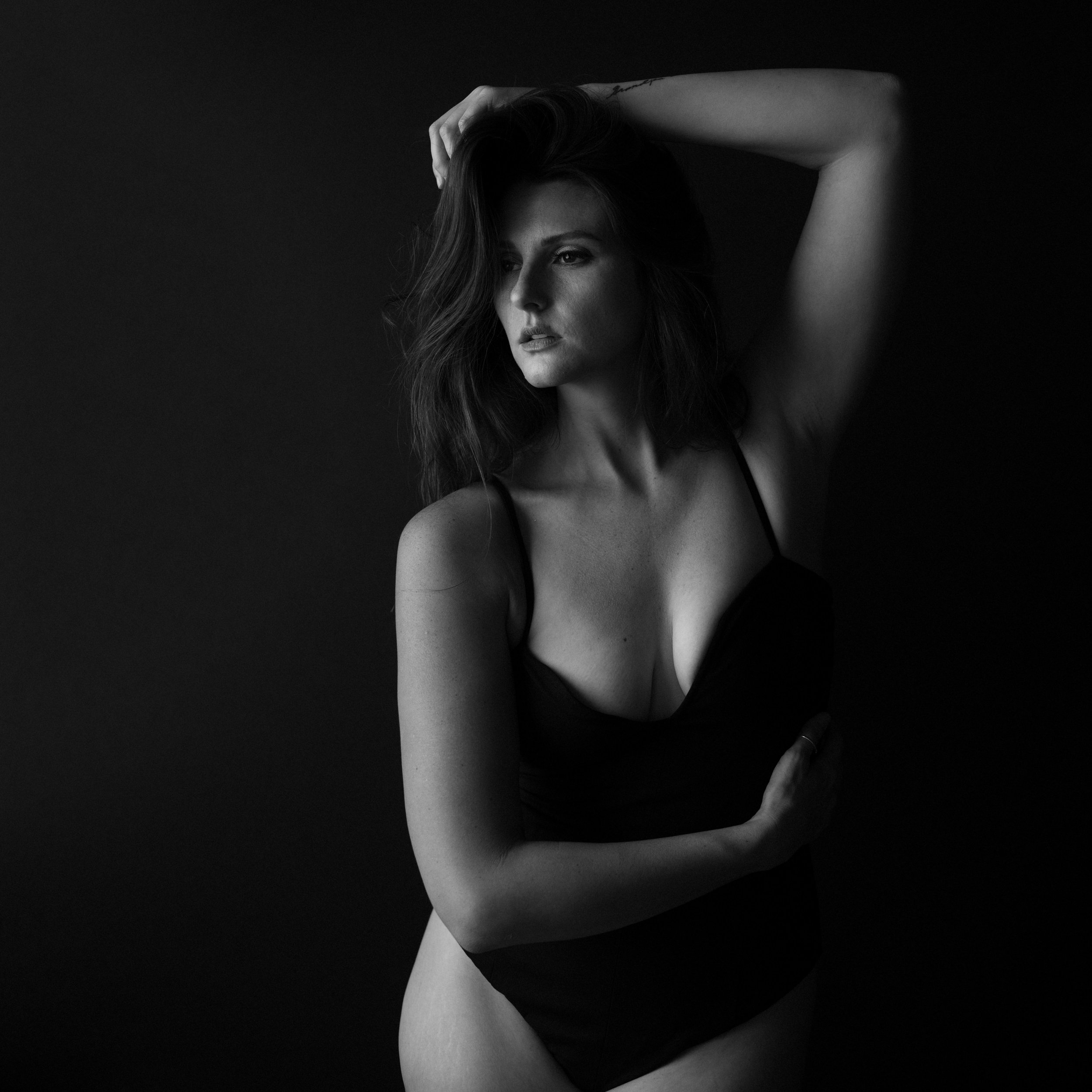 Black and white photo of a woman with wavy hair in a black dress, standing against a dark background, with one arm raised and hand in her hair, gazing thoughtfully.