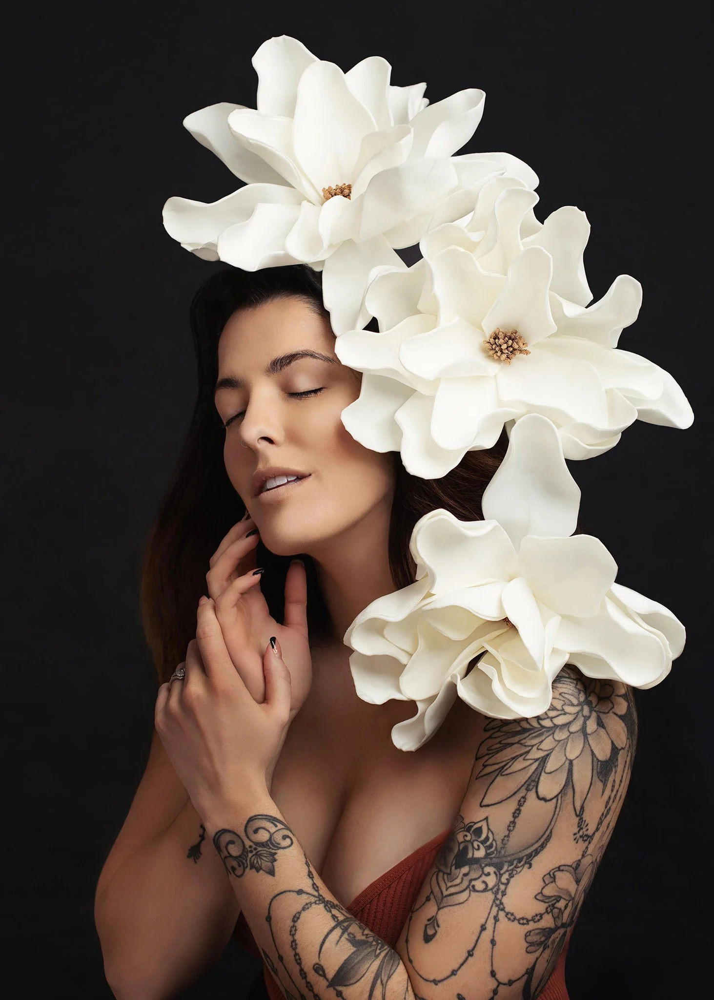 A woman with dark hair and tattoos on her arms, with her eyes closed, touching her face, adorned with large white flowers around her head against a black background.