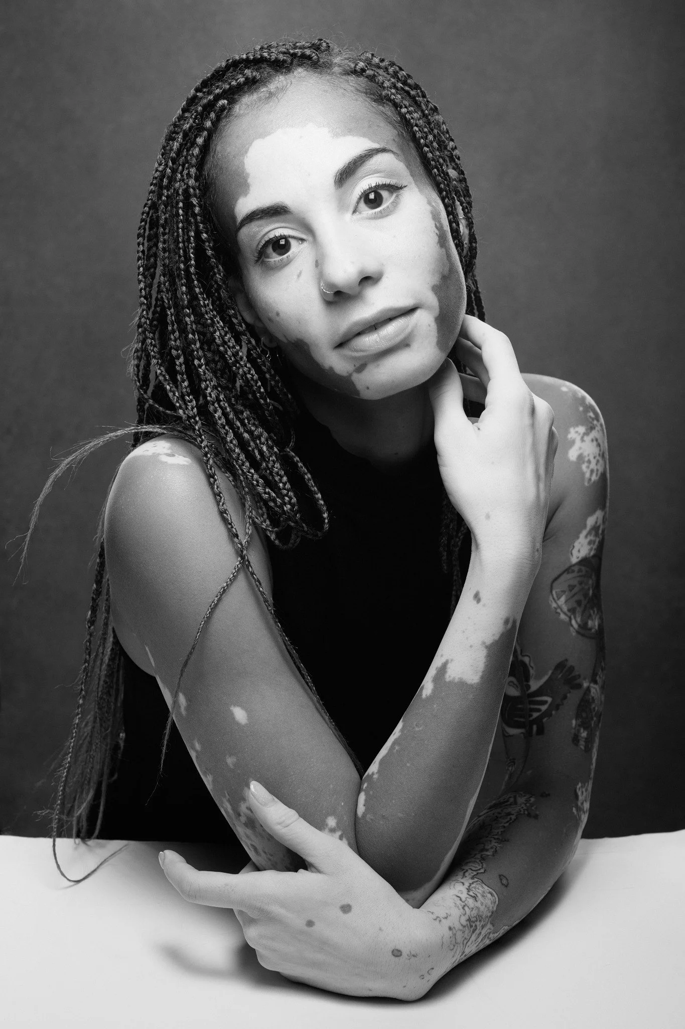 A young woman with braided hair and tattoos on her arms, sitting at a table, touching her neck with her right hand, looking at the camera, in a black and white portrait.
