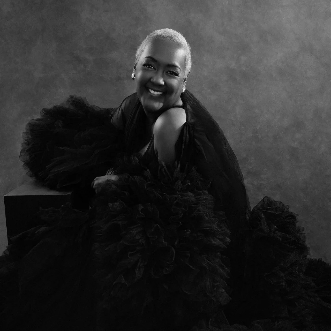 Black and white portrait of a smiling woman with short, platinum blonde hair, wearing a voluminous black dress with ruffled layers, sitting and leaning on her left arm against a textured background.