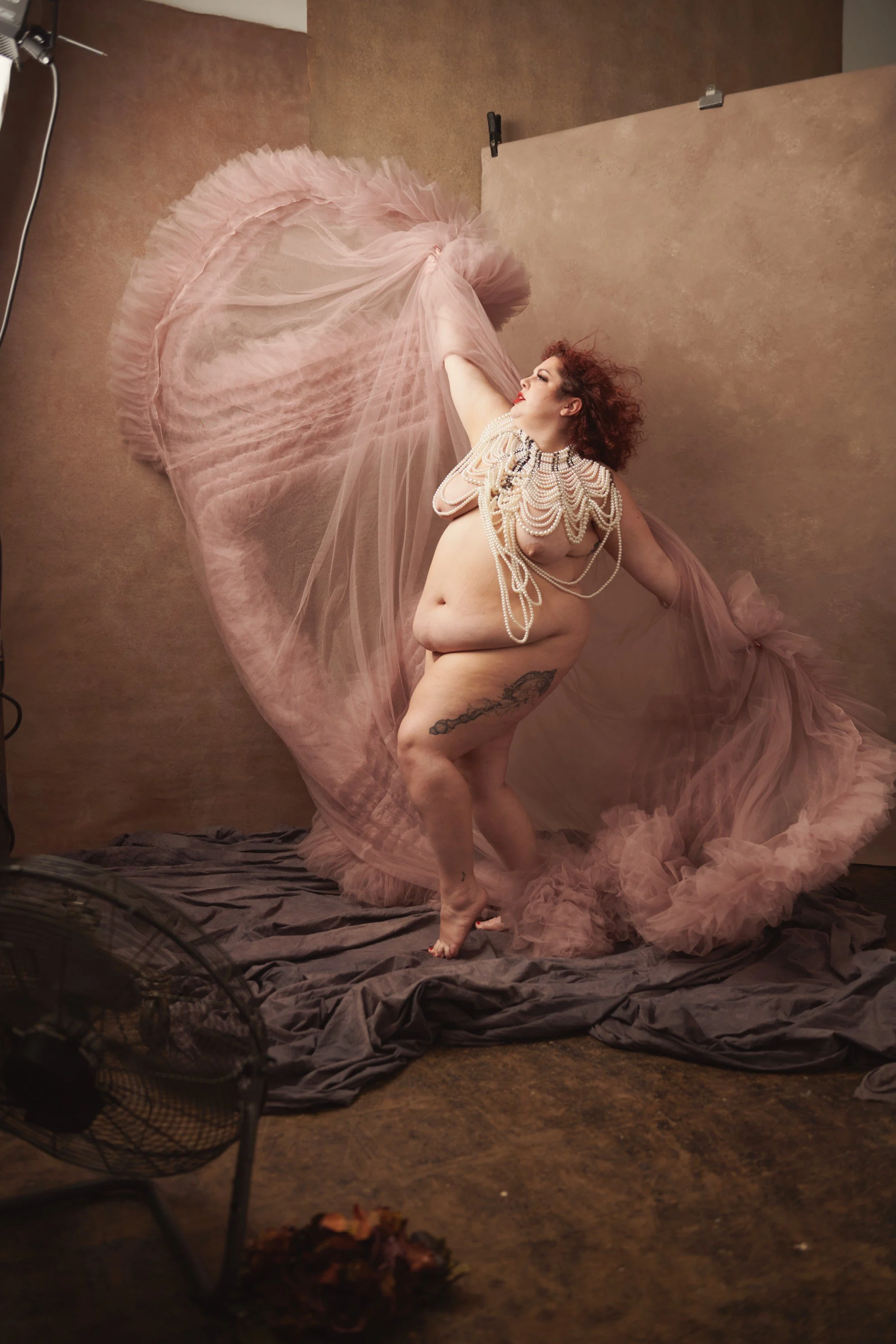 A woman posed in a photography studio, surrounded by pink tulle fabric and wearing layered pearl necklaces.