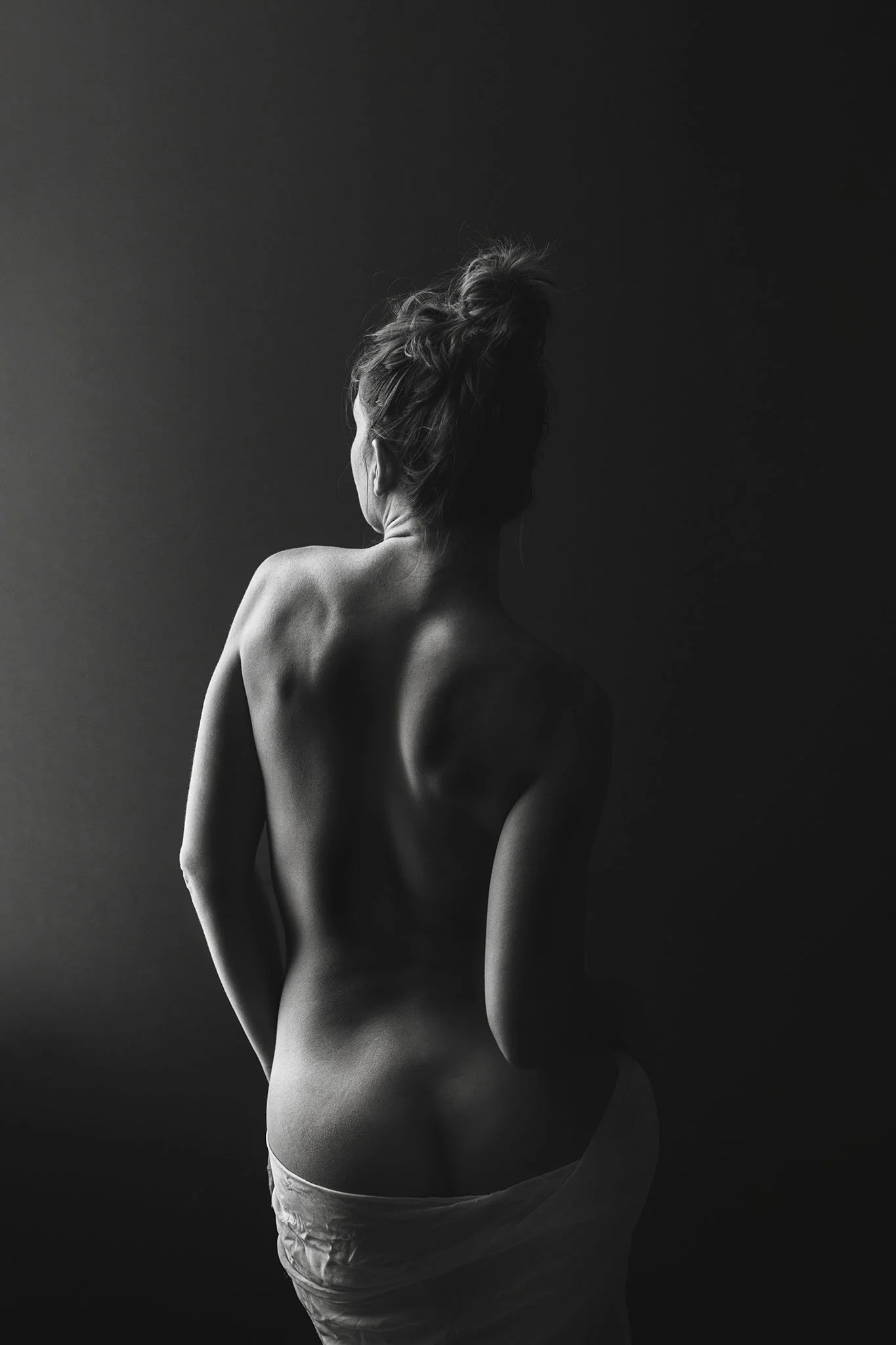 A black and white photo of a woman viewed from the back, with her hair tied up, partially covered by a cloth around her hips, and illuminated with soft light highlighting her back and shoulder contours.