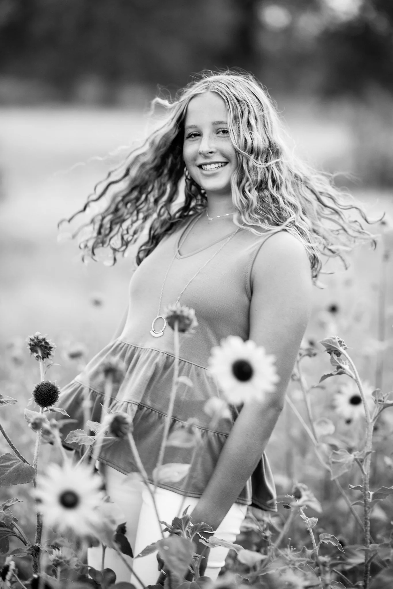A young woman with curly hair smiling and standing among tall sunflowers in a black-and-white photo.