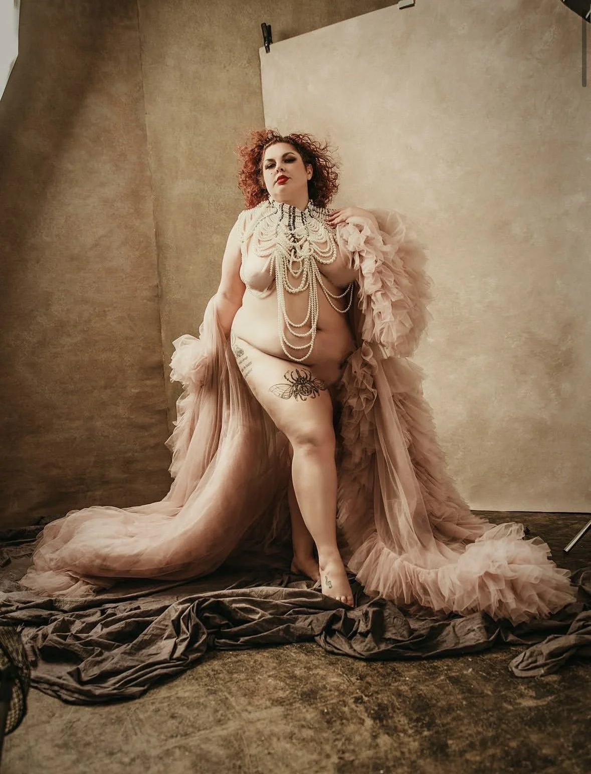 A woman with curly red hair and tattoos posing topless with a long, flowing pink tulle gown and layered pearl necklaces in a photo studio with a beige backdrop and fabric on the floor.