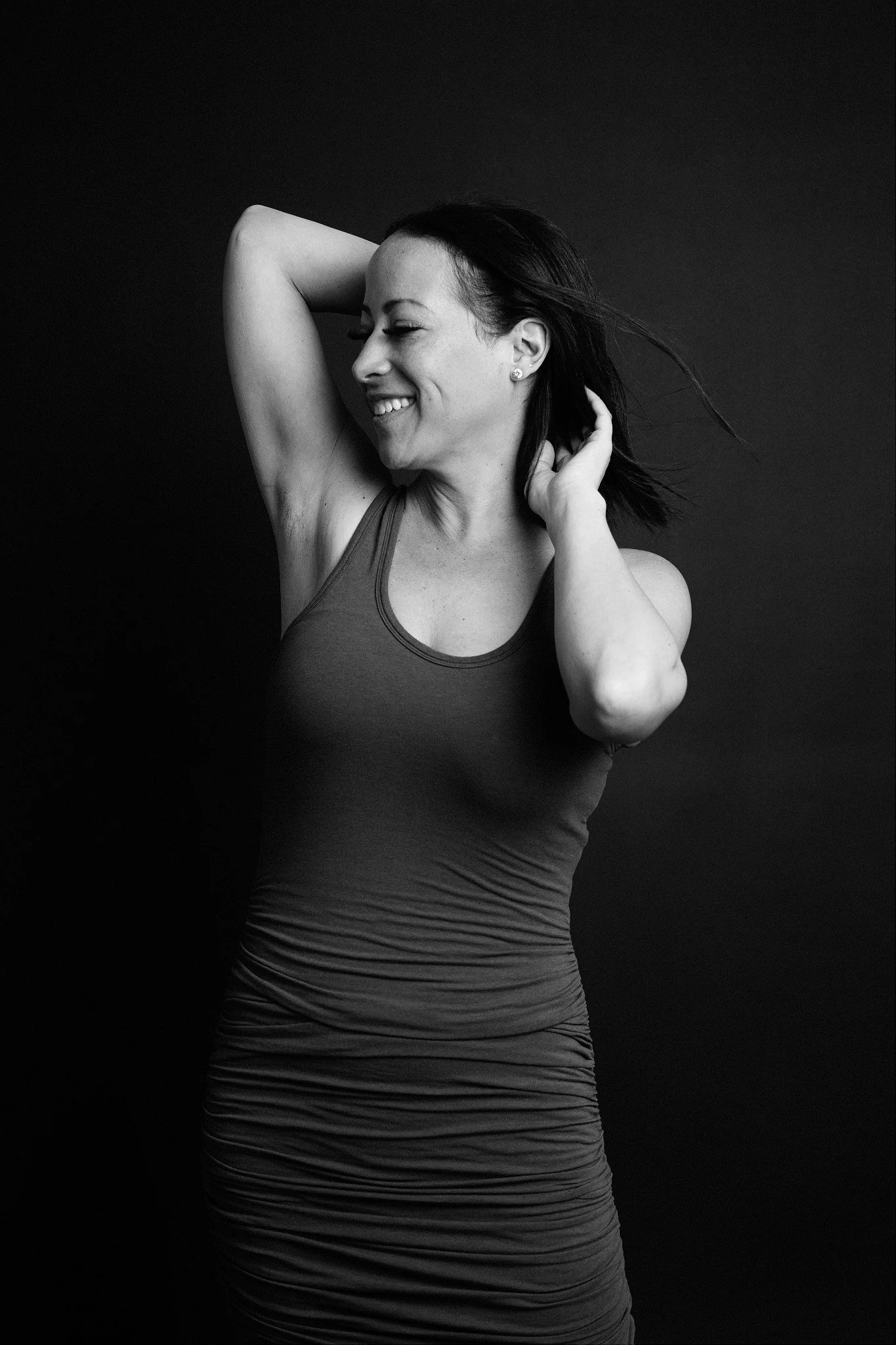 Black and white photo of a woman smiling with her eyes closed, wearing a sleeveless top, with one arm raised and her hand behind her head, against a plain dark background.