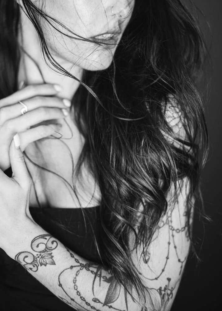 Black and white photo of a woman with long, wavy hair, tattoos on her arm, and a ring on her finger, with her hand on her neck.