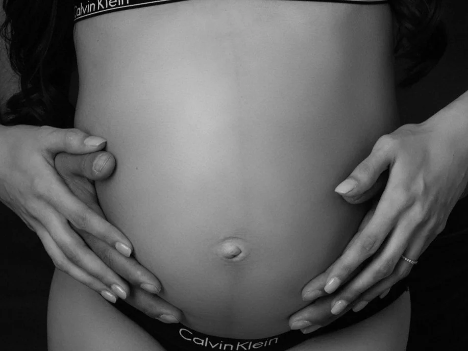 A pregnant woman with her hands gently cradling her belly, wearing Calvin Klein underwear, black and white photo.