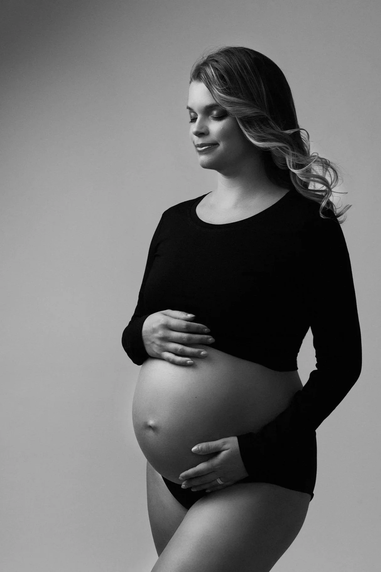 Black and white photo of a pregnant woman with long hair, smiling, wearing a black long sleeve crop top, holding her baby bump