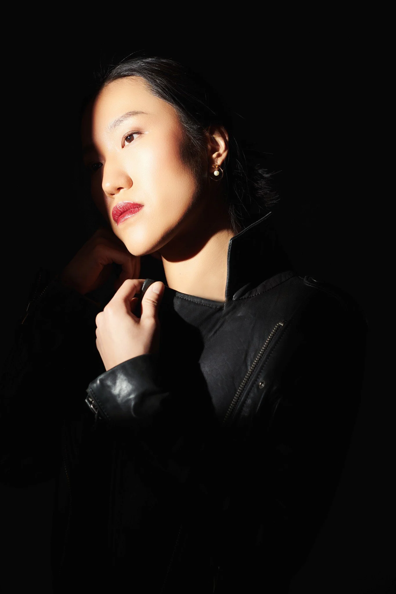 A woman with dark hair and earrings, wearing a black leather jacket, looking to the side with her hand near her chin, against a dark background.