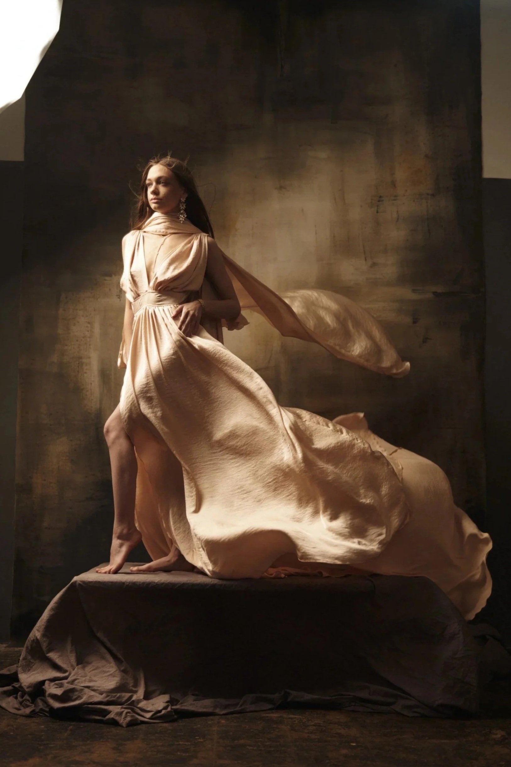 A woman in a flowing, elegant, beige gown posing on a stone pedestal against a dark, textured background.