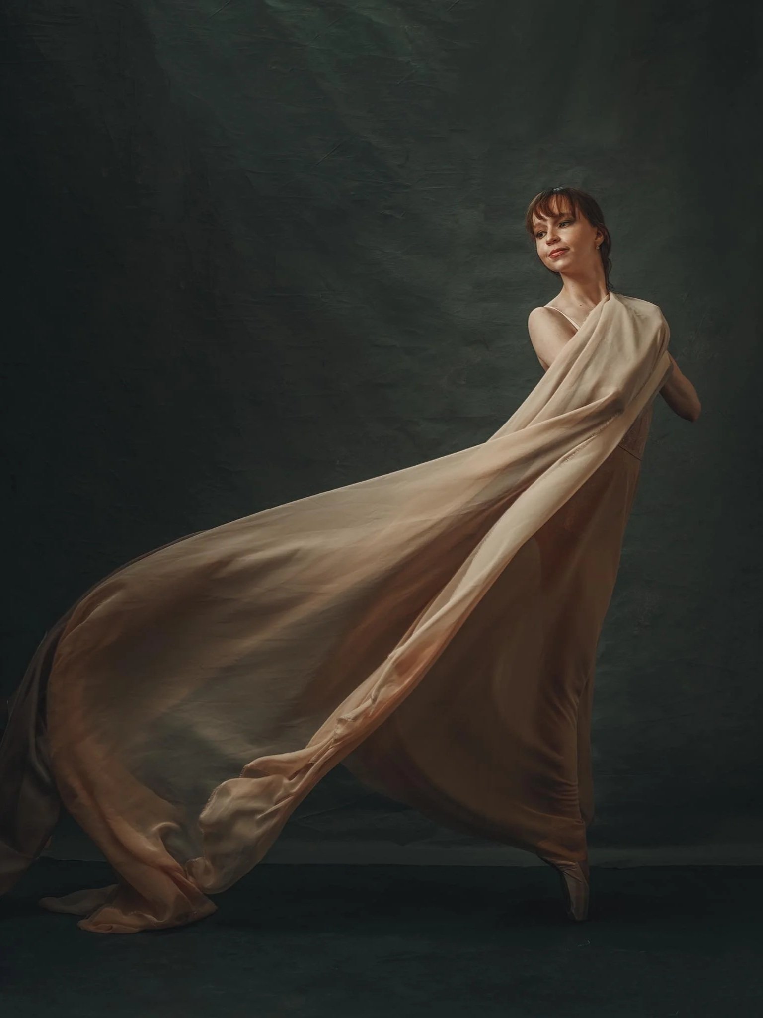 A woman standing in front of a dark background, wrapped in a flowing, beige fabric that trails behind her.