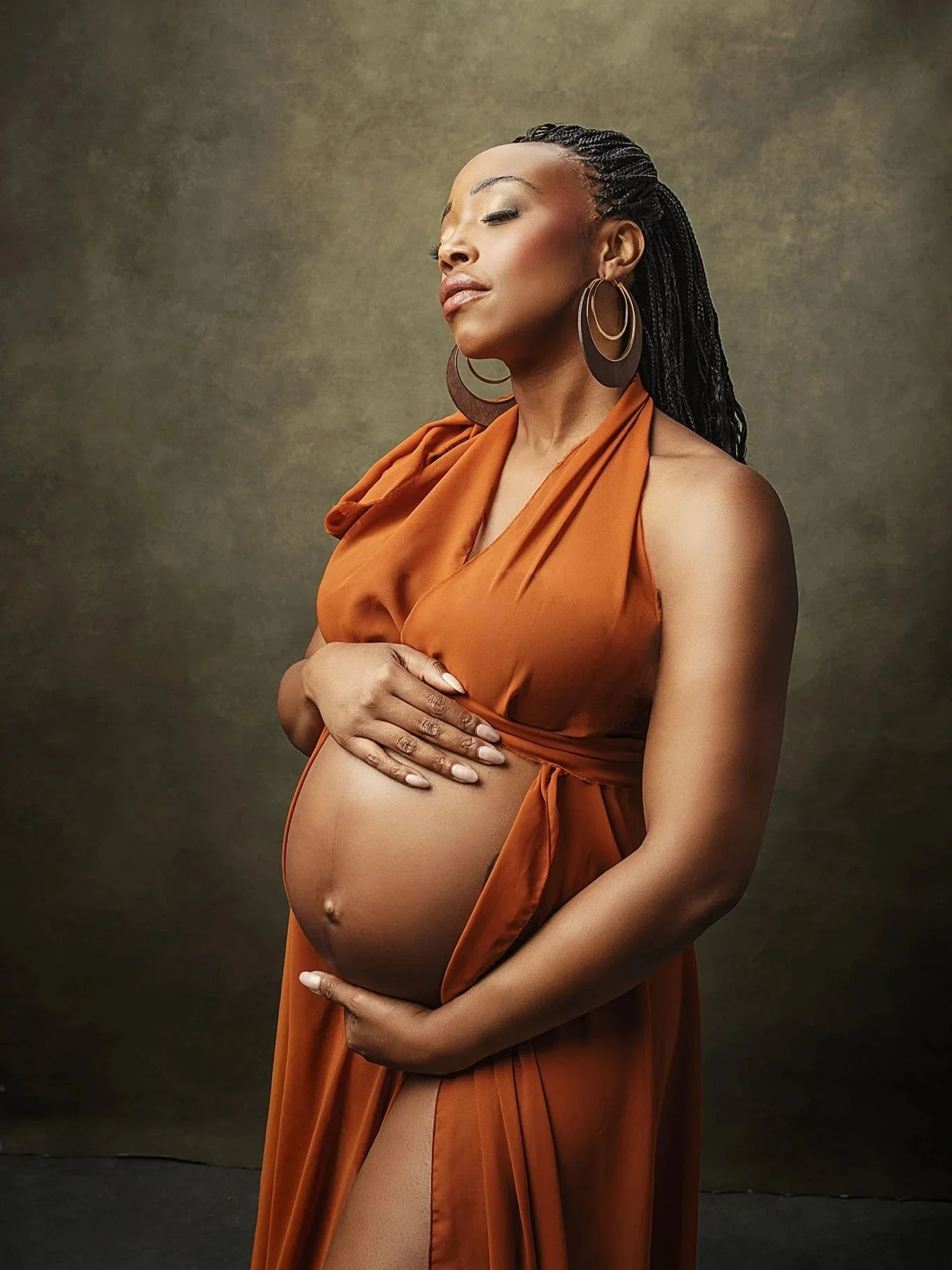 A pregnant woman with braided hair wearing a rust-colored dress and large hoop earrings, holding her belly with one hand and standing against a plain background.
