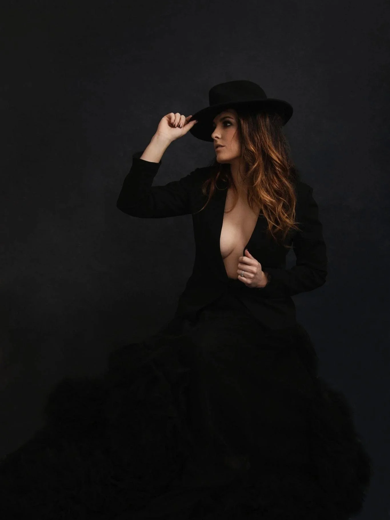 A woman wearing a black hat and a black blazer with a deep neckline, sitting against a dark background.