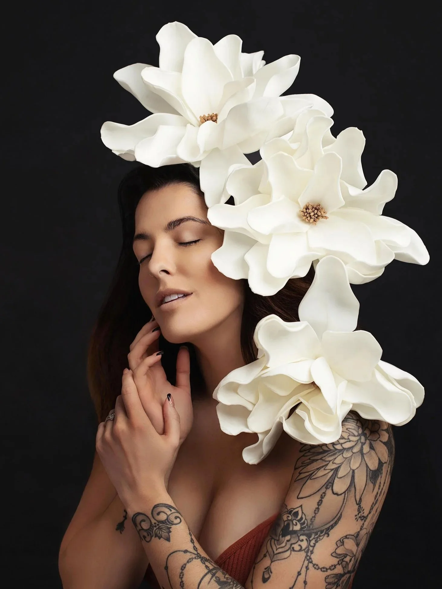 A woman with closed eyes and tattoos on her arms and chest, holding her hands near her face, with large white flowers on her head and shoulder against a black background.