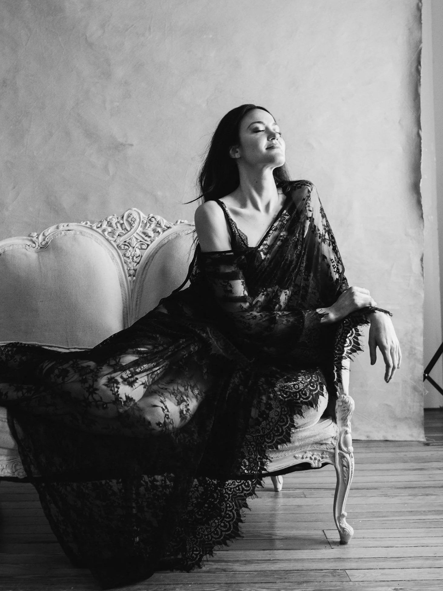 A woman in a sheer black lace dress sitting on an ornate vintage sofa with a plain wall background, black and white photo.