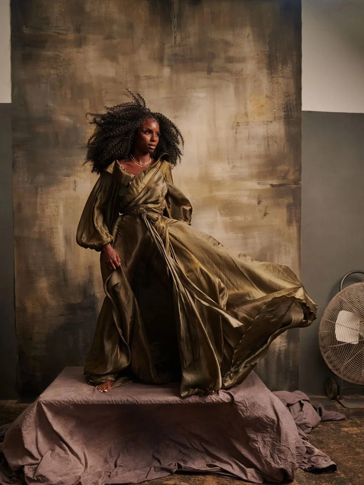 A woman with curly hair standing on a fabric-covered platform in front of a textured neutral-toned backdrop, wearing a gold satin gown with voluminous sleeves and a matching sash, with a fan visible on the right side.