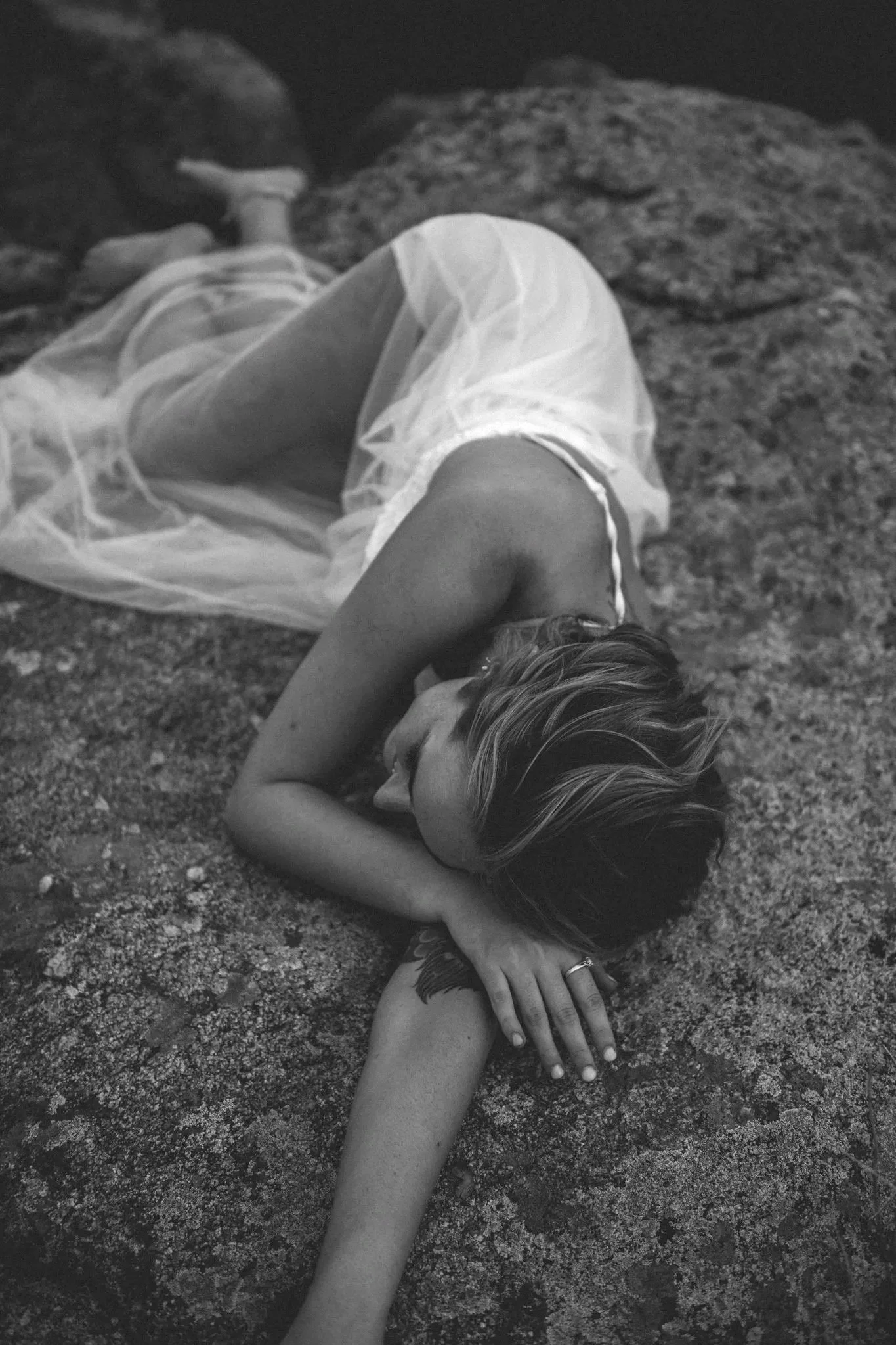 A woman lying on a rocky surface with her arm extended, wearing a sheer skirt and a sleeveless top, with her head resting on her arm and her eyes closed.