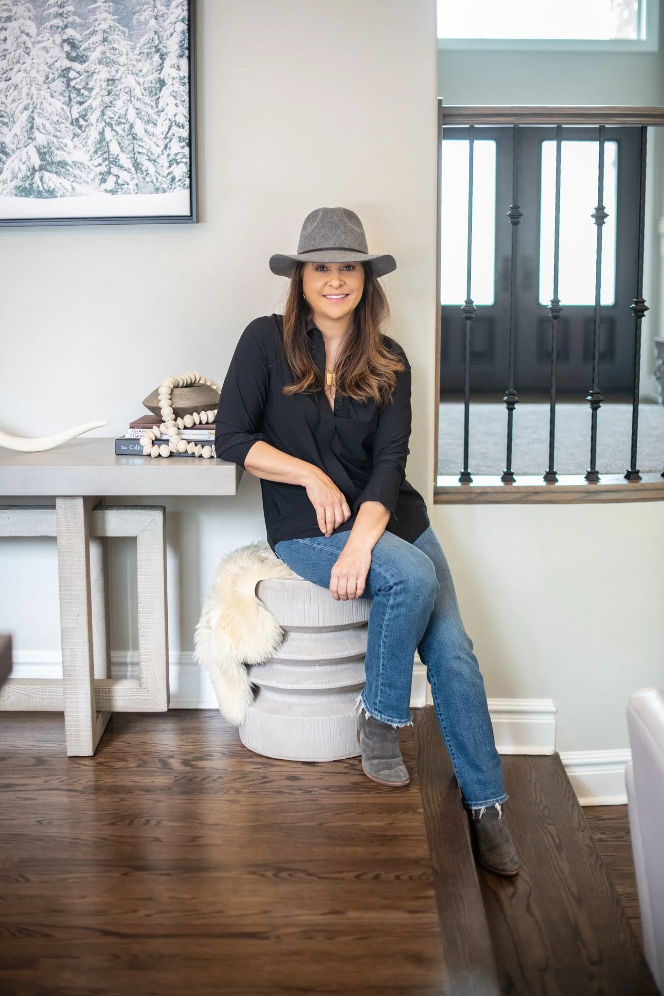 A woman wearing a gray hat, black shirt, blue jeans, and gray ankle boots, sitting on a white stool with a furry throw, posing indoors near a wall with a window and a side table with books and decorative objects.