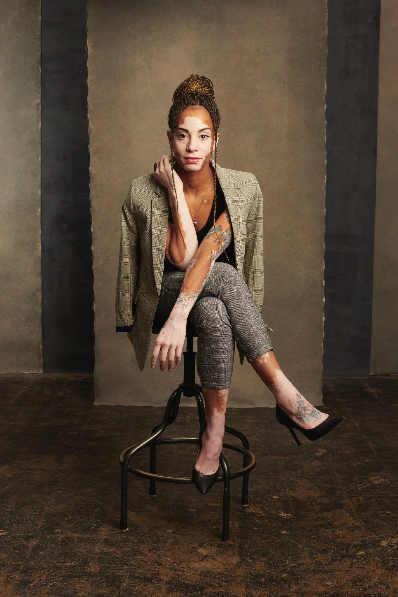A woman with tattoos and braids, dressed in a plaid blazer, black top, and plaid pants, sitting on a stool in front of textured brown and black background.